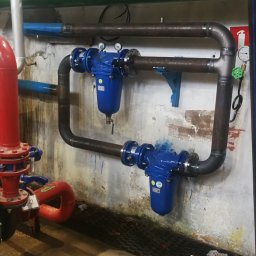 MATEUSZ BARAŃSKI KREWICUSTOM - Instalacja przemysłowa z rur stalowych i niebieskich filtrów, widoczne połączenia spawane, zawory i manometry, tło stanowi ściana z widocznymi śladami użytkowania.