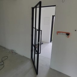 MATEUSZ BARAŃSKI KREWICUSTOM - Częściowo otwarta, czarna, metalowa rama drzwi z szybami w minimalistycznym, surowym wnętrzu w trakcie wykończenia.