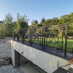 Nowoczesna, czarna balustrada z geometrycznym wzorem na tarasie z widokiem na zadbany ogród i mur oporowy z gabionów.