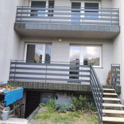 MATEUSZ BARAŃSKI KREWICUSTOM - Dwukondygnacyjny budynek z balkonami zabezpieczonymi poziomymi, szarymi barierkami. Schody z drewnianymi stopniami prowadzą na balkon. Widoczne narzędzia ogrodnicze.