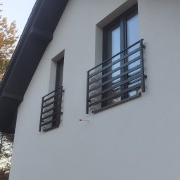 MATEUSZ BARAŃSKI KREWICUSTOM - Elewacja domu z dwoma oknami balkonowymi zabezpieczonymi czarnymi, metalowymi balustradami o minimalistycznym designie. Widoczny fragment dachu i drzewa w tle.