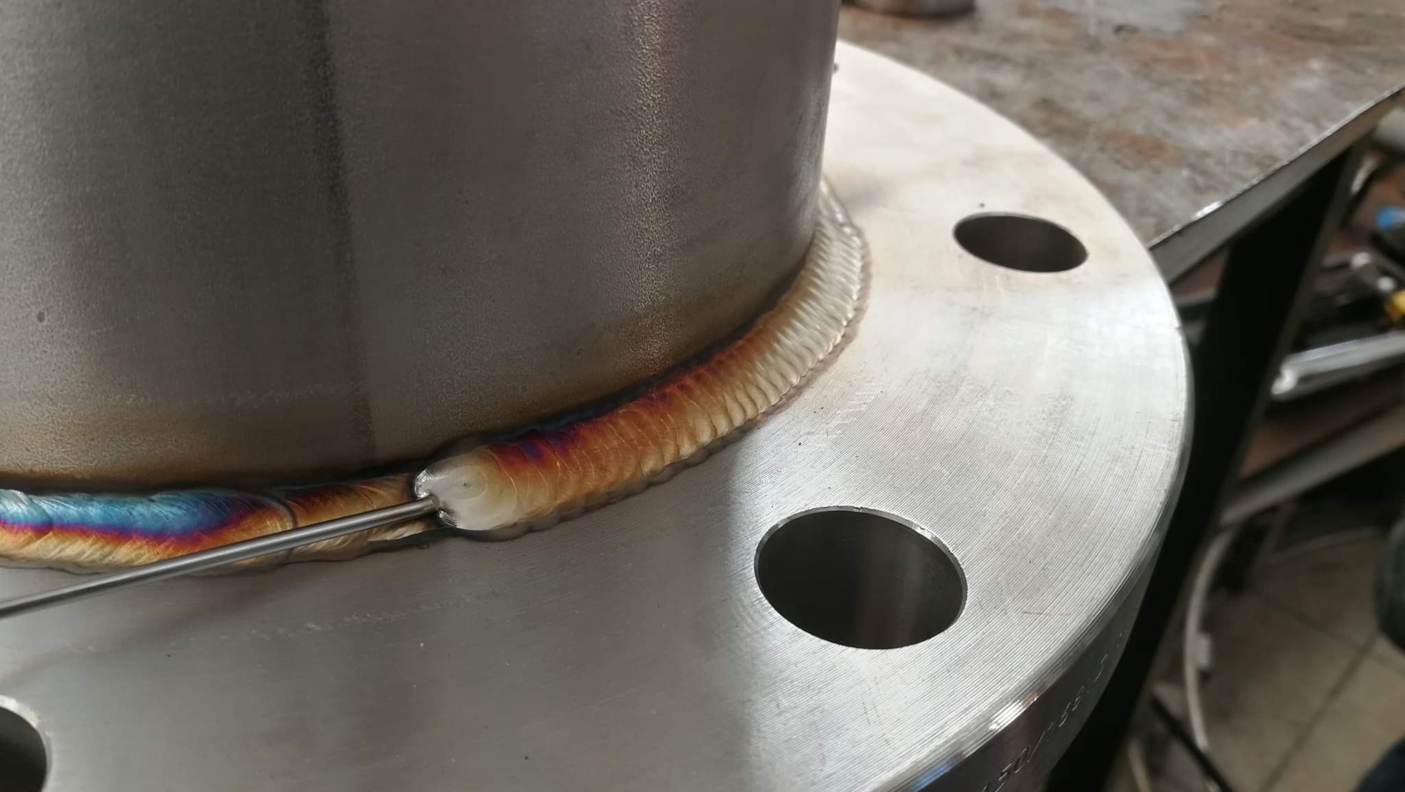 Detal spoiny na rurze stalowej z kołnierzem, widoczne tęczowe przebarwienia i drut spawalniczy.
