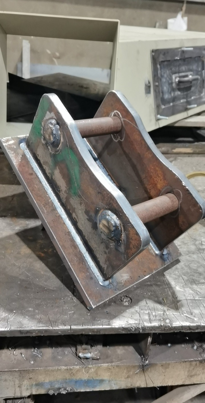 Metalowy uchwyt z dwiema tulejami, spawany do podstawy, widoczne ślady spawania i obróbki, na tle warsztatu.