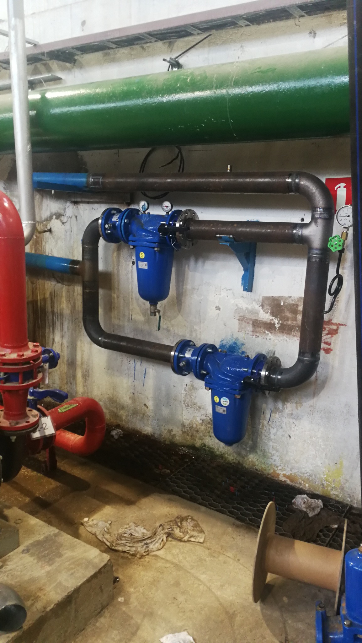 Instalacja przemysłowa z rur stalowych i niebieskich filtrów, widoczne połączenia spawane, zawory i manometry, tło stanowi ściana z widocznymi śladami użytkowania.