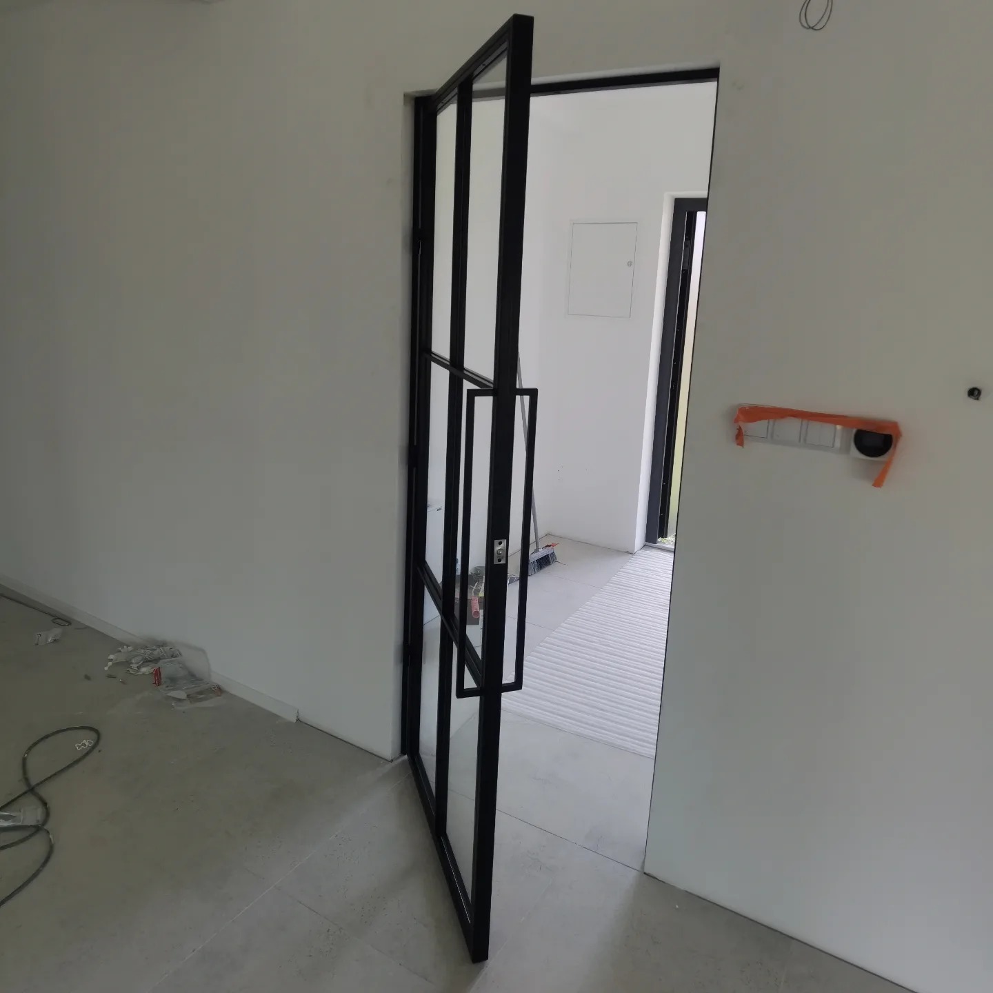 Częściowo otwarta, czarna, metalowa rama drzwi z szybami w minimalistycznym, surowym wnętrzu w trakcie wykończenia.