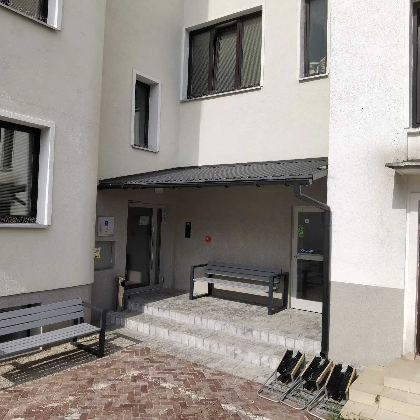 Zadaszone wejście do budynku z szarymi ławkami, schodami z jasnych płytek i stojakiem na szczotki do butów na ceglanej posadzce.