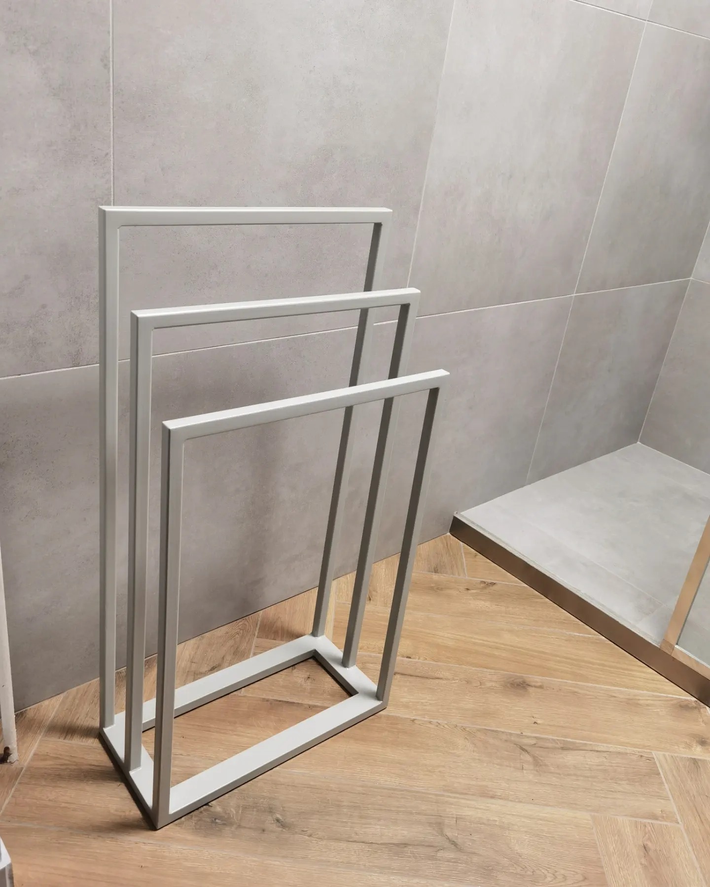 Metalowy, minimalistyczny stojak na ręczniki w łazience z szarymi płytkami i drewnopodobną podłogą ułożoną w jodełkę.