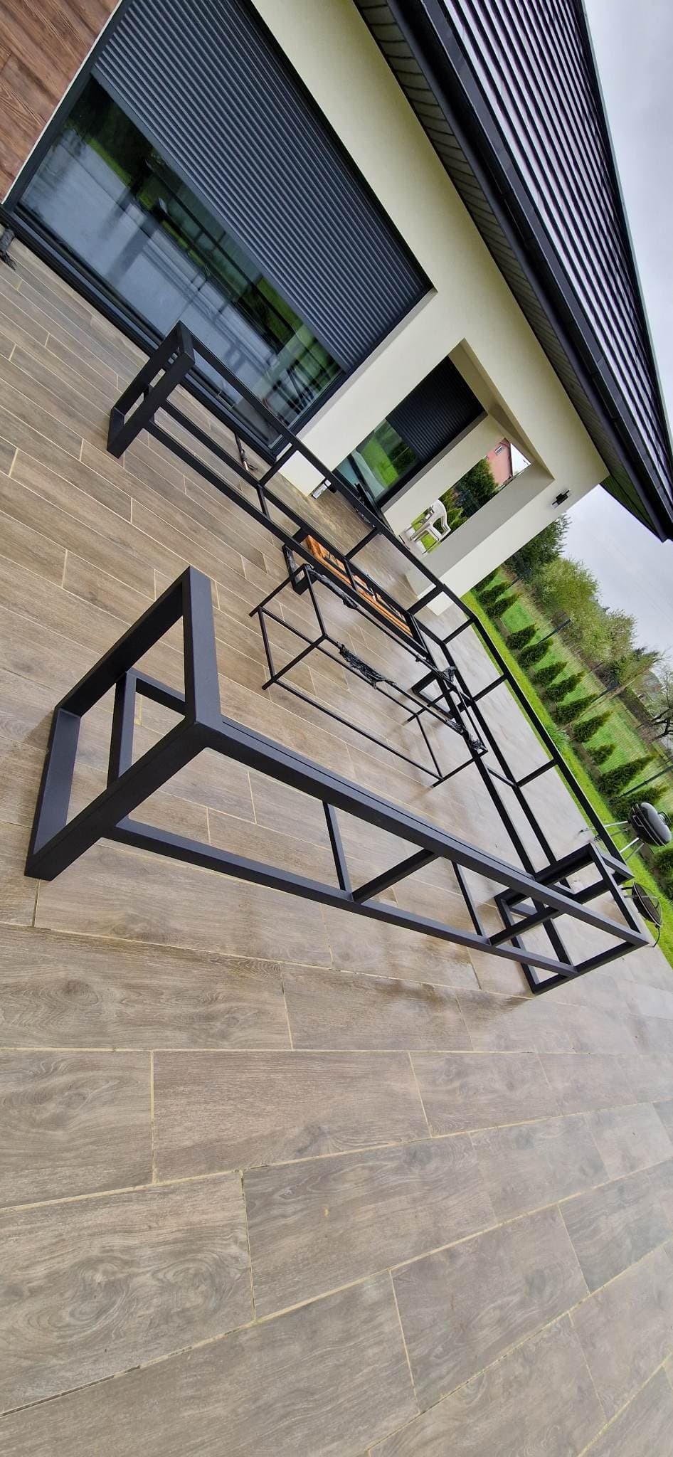 Czarne, metalowe ramy konstrukcji pergoli lub zadaszenia tarasu, rozłożone na drewnopodobnych płytkach tarasowych przed nowoczesnym domem z roletami zewnętrznymi.