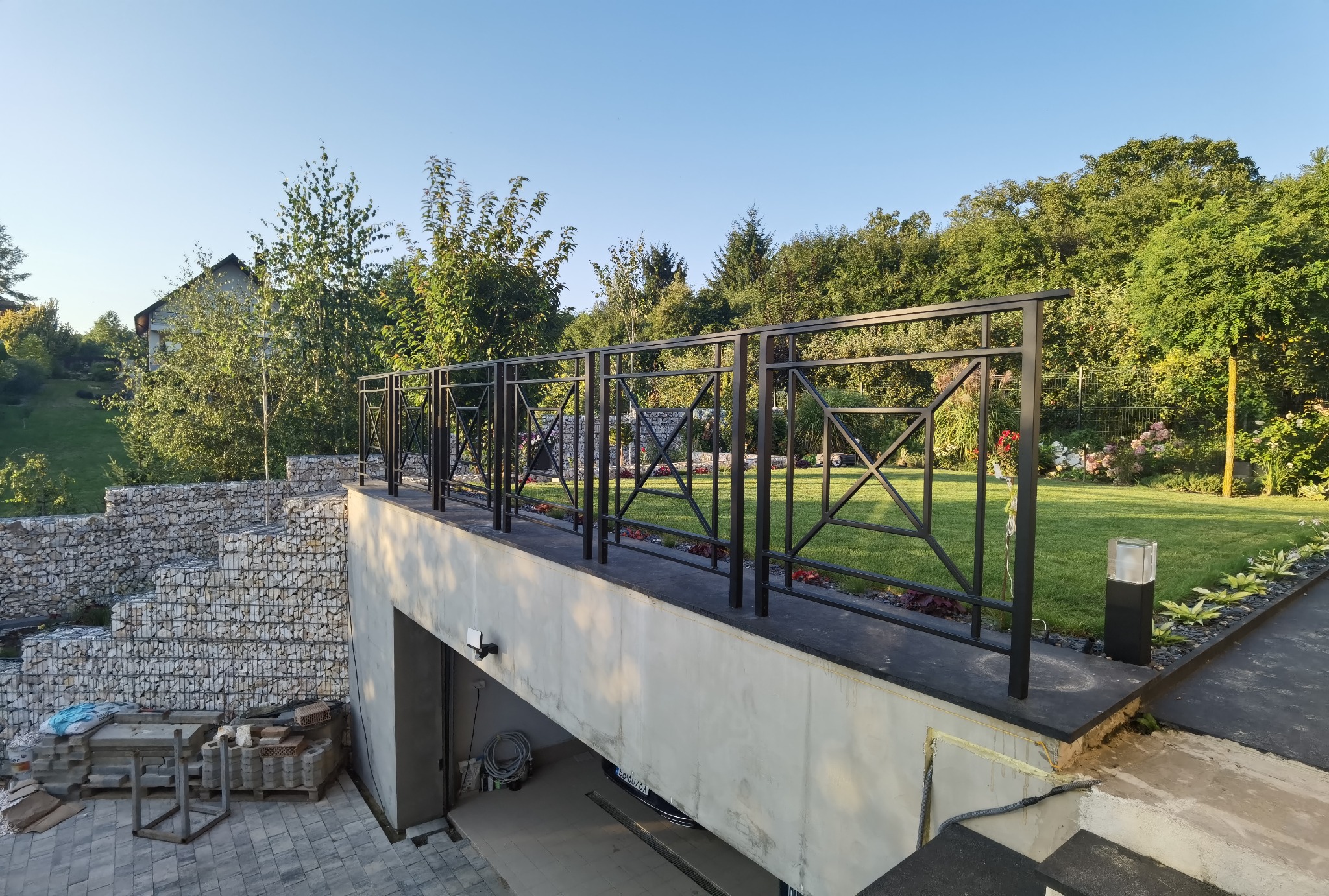 Nowoczesna, czarna balustrada z geometrycznym wzorem na tarasie z widokiem na zadbany ogród i mur oporowy z gabionów.