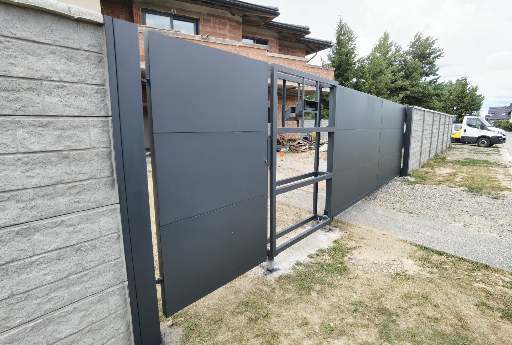 Nowoczesna, przesuwna brama wjazdowa z metalu, częściowo otwarta, widoczne elementy konstrukcyjne i betonowe słupki ogrodzeniowe w tle, teren w trakcie zagospodarowywania.