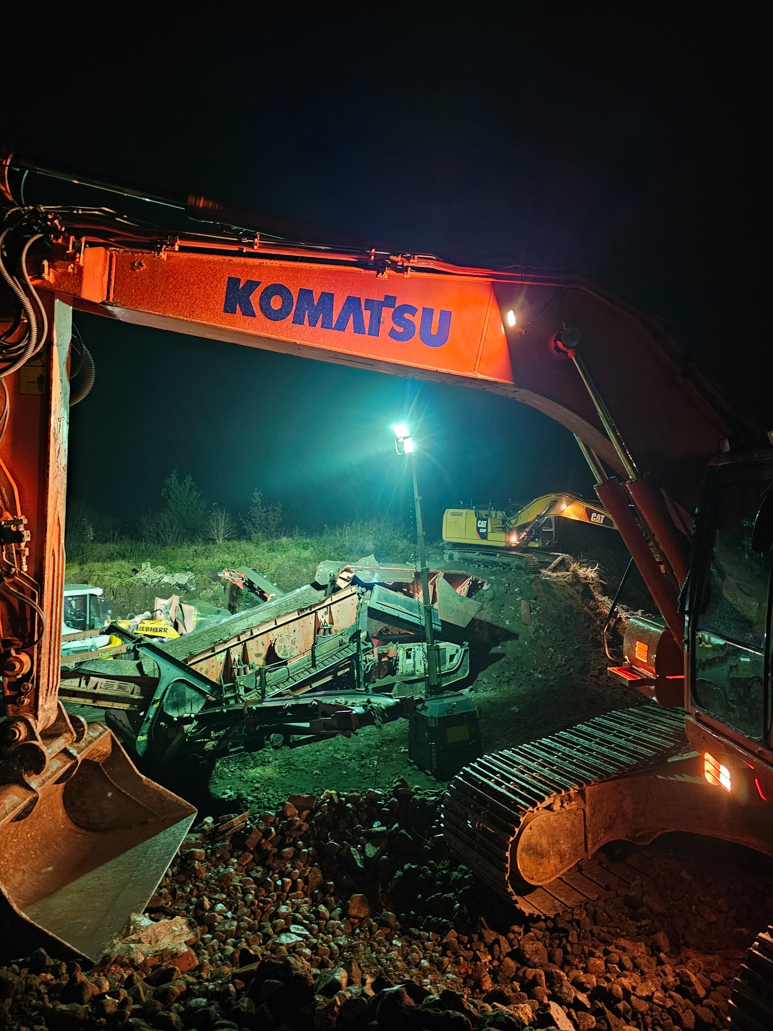 Nocne ujęcie placu budowy z pomarańczowym koparką Komatsu na pierwszym planie, oświetloną lampą halogenową, w tle widoczna żółta koparka CAT i przesiewacz do gruzu, teren usypany kamieniami.