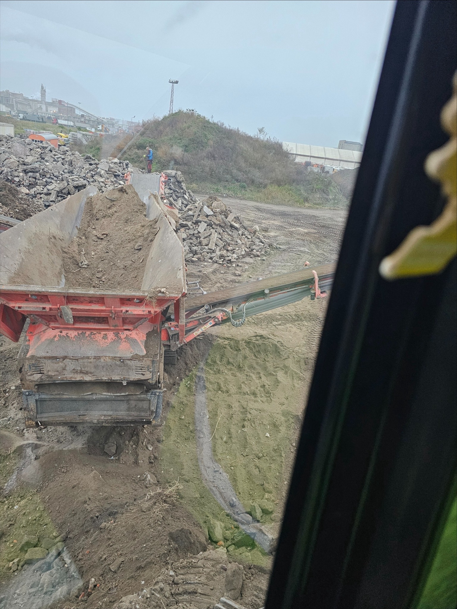 Widok z kabiny na maszynę przesiewającą gruz na placu budowy z pracownikiem w oddali, w tle zabudowania przemysłowe i wzgórze.