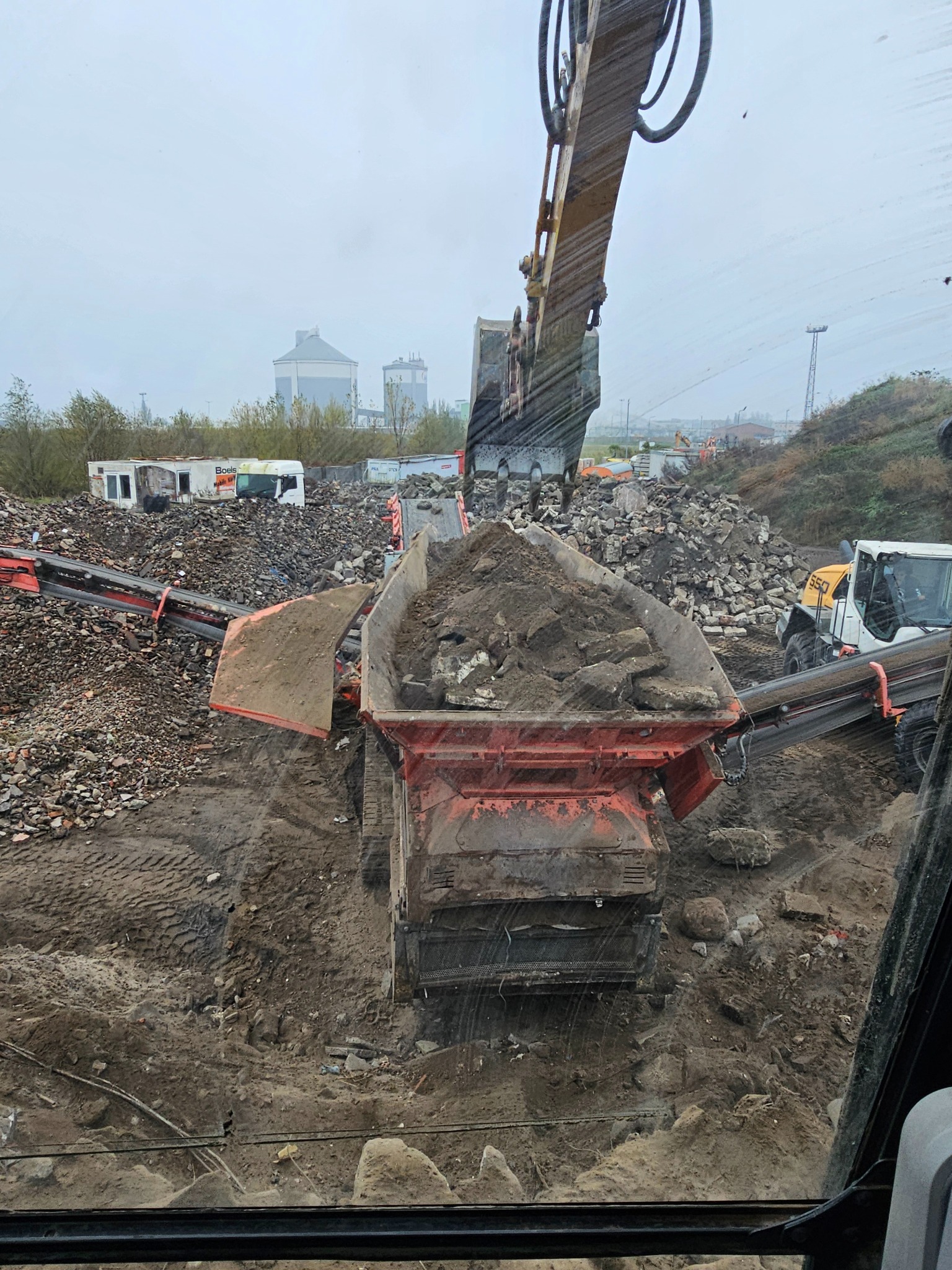 Praca maszyny budowlanej, prawdopodobnie kruszarki, podczas przetwarzania gruzu w Kobylinie. Widok z kabiny operatora na załadunek materiału do kruszarki. W tle składowisko gruzu i zabudowania.