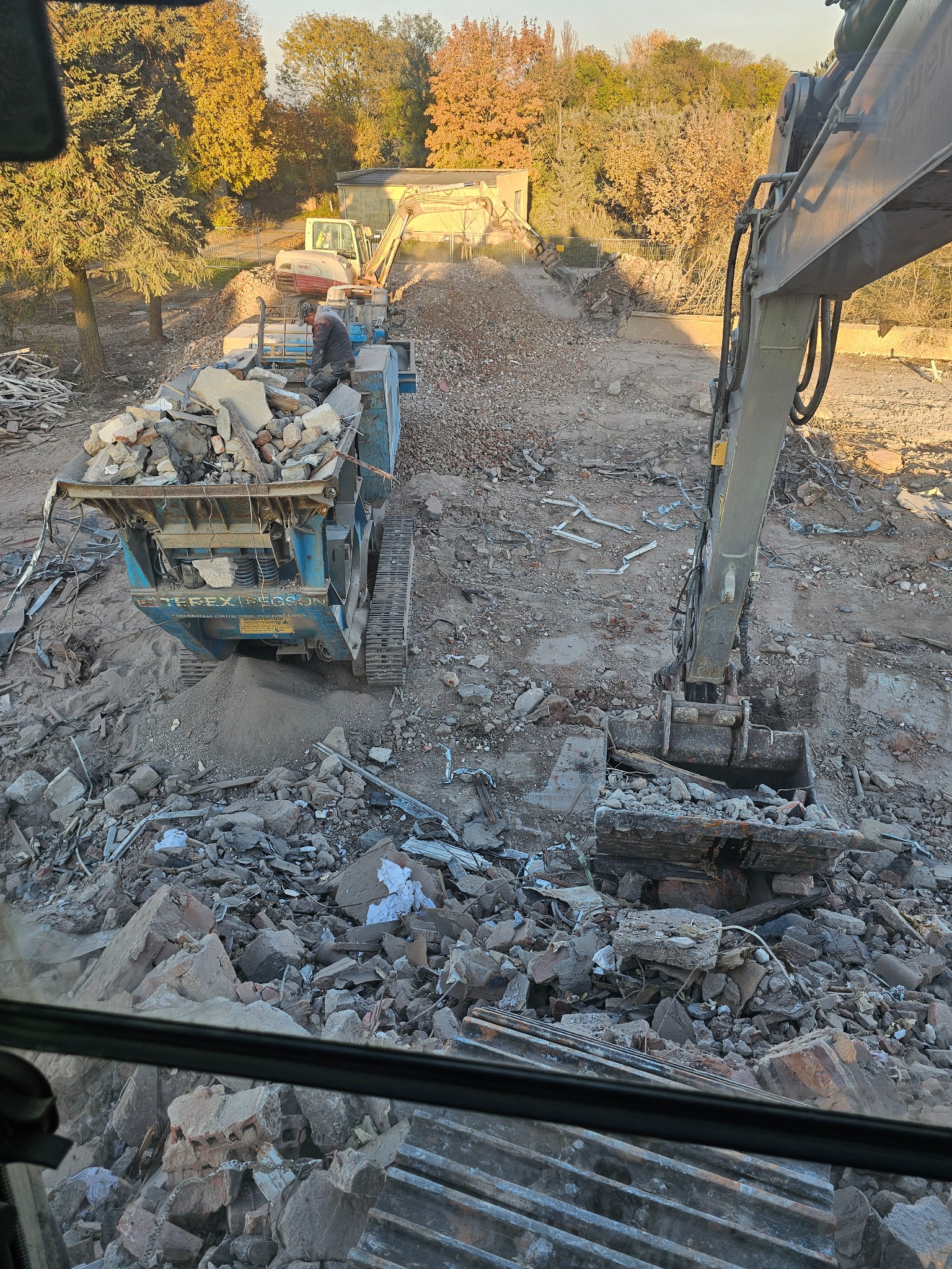 Demontaż konstrukcji budowlanej: koparka załadowuje gruz do przesiewacza mobilnego, w tle widoczna inna maszyna i drzewa w jesiennej scenerii.