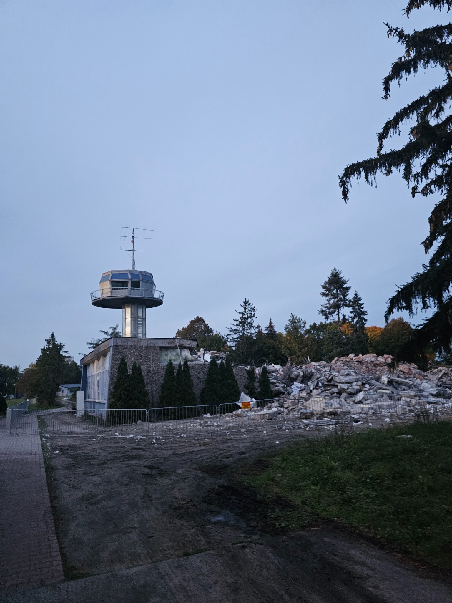 Budynek z wieżą obserwacyjną częściowo zburzony, widoczne gruzy i zabezpieczenia, w tle drzewa i pochmurne niebo.