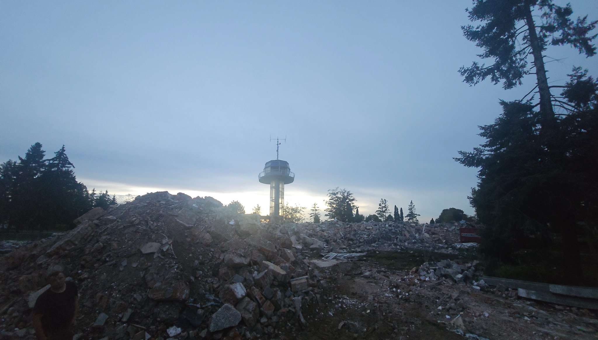 Góra gruzu po rozbiórce budynku, z widoczną wieżą obserwacyjną w tle, sfotografowana o zmierzchu.