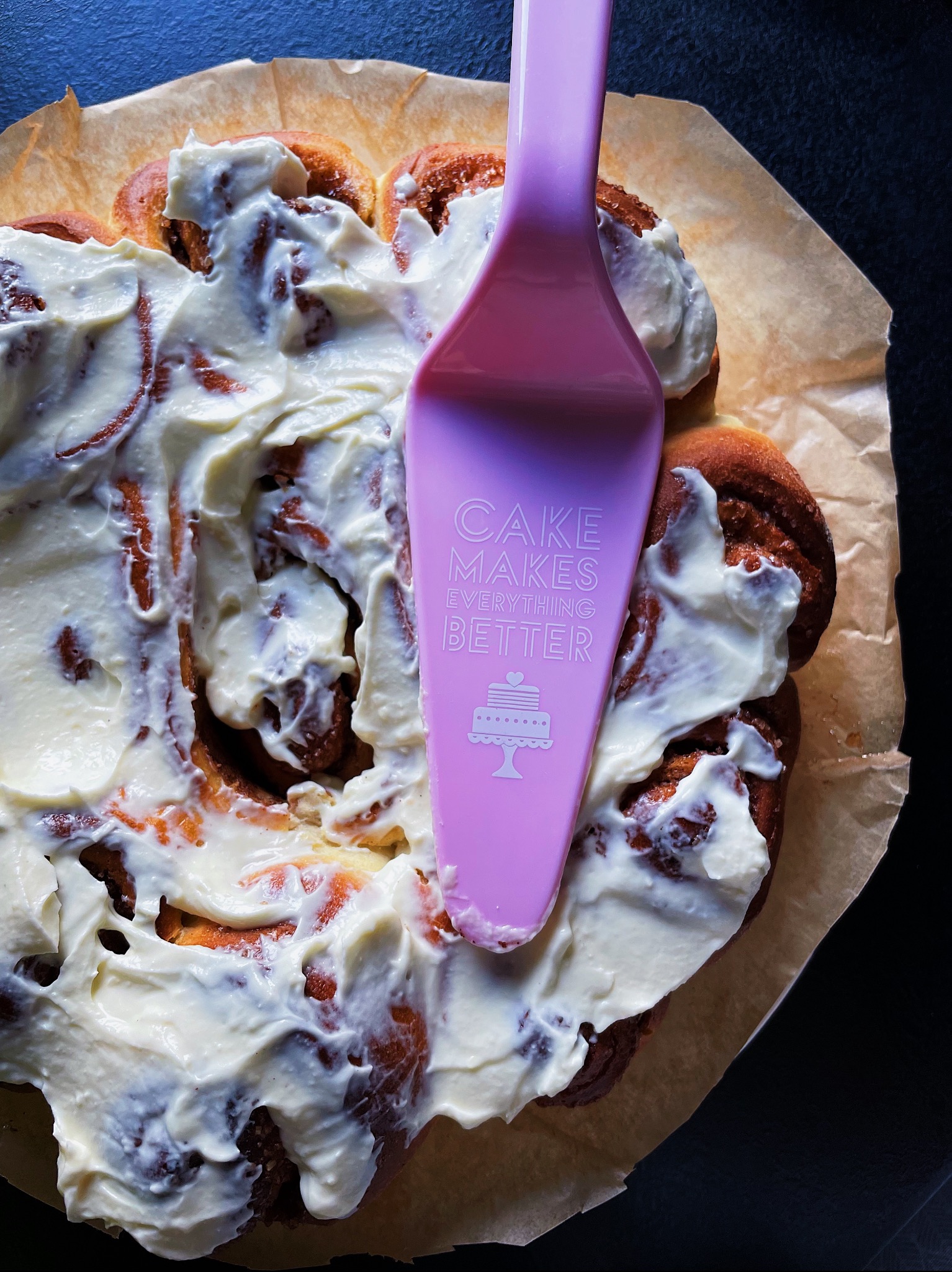 Cynamonki na papierze do pieczenia, pokryte kremowym lukrem, z różową łopatką do ciasta z napisem 'CAKE MAKES EVERYTHING BETTER'.