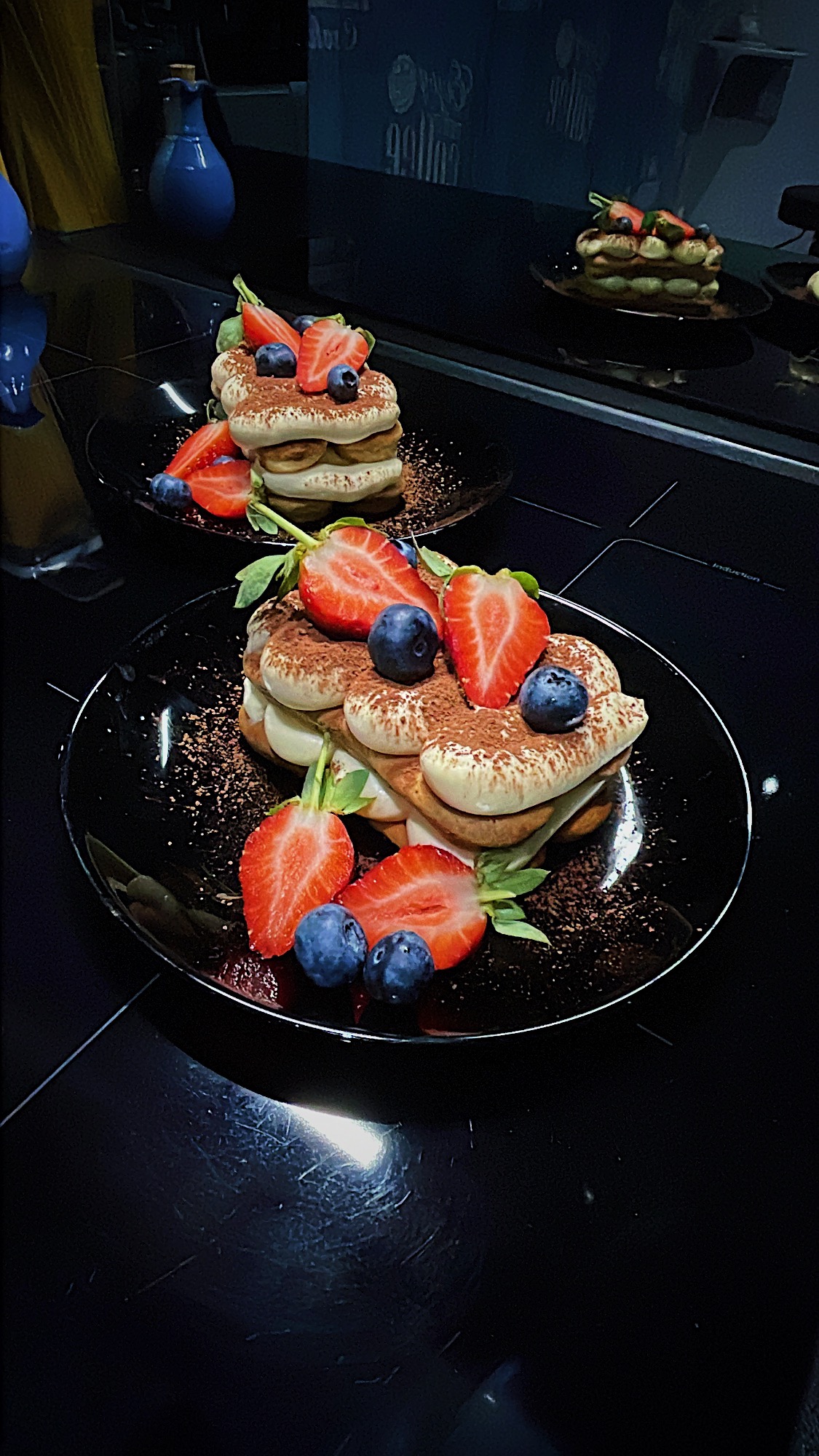 Dwa talerze z deserem tiramisu udekorowanym świeżymi truskawkami i borówkami, posypane kakao, na błyszczącym czarnym blacie.