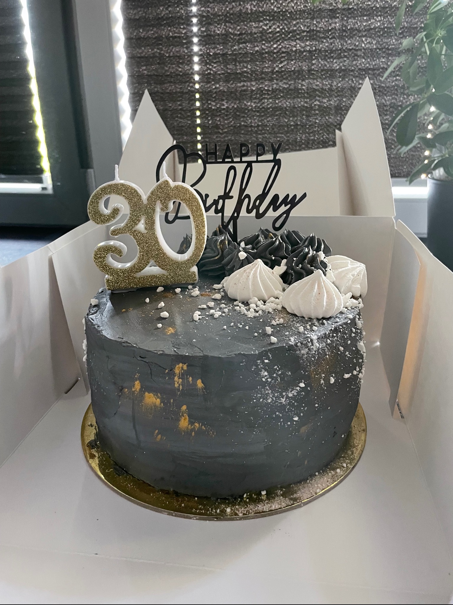 Tort urodzinowy w odcieniach szarości z dekoracjami z bezy i napisem 'Happy Birthday' oraz świeczką w kształcie liczby 30, umieszczony w białym pudełku.