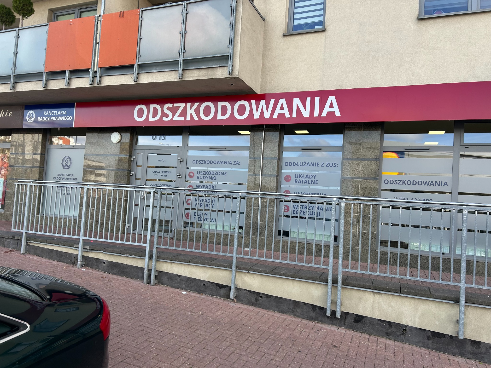 Fasada budynku z szyldem 'Odszkodowania', widoczny napis 'Kancelaria Radcy Prawnego' oraz informacje o odszkodowaniach za uszkodzone budynki i wypadki przy pracy.