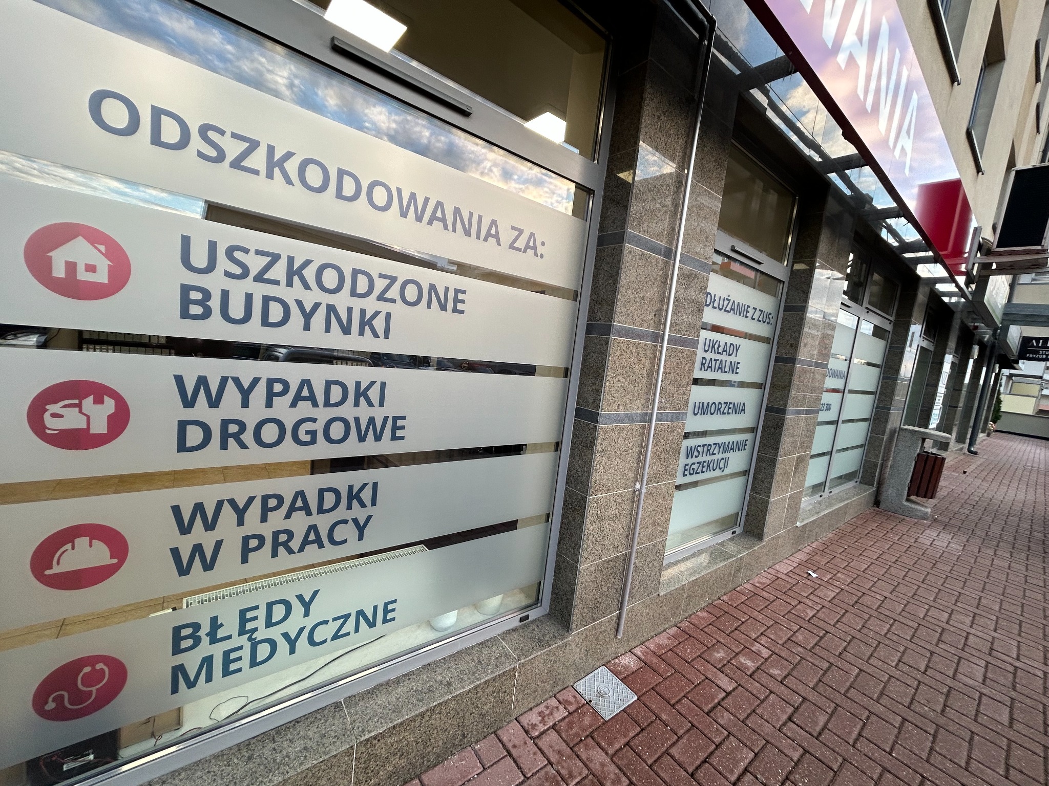 Zewnętrzny widok na okno biura z napisem 'Odszkodowania za: Uszkodzone Budynki, Wypadki Drogowe, Wypadki w Pracy, Błędy Medyczne' oraz 'Dłużanie z ZUS: Układy Ratalne, Umorzenia, Wstrzymanie...