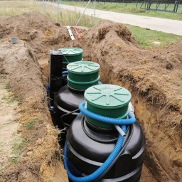 Domowe-oczyszczlanie - Instalacja trzech zbiorników z zielonymi pokrywami w wykopie, widoczne niebieskie rury karbowane i czarna folia ochronna, ziemia wokół, w tle ogrodzenie z siatki i droga gruntowa.