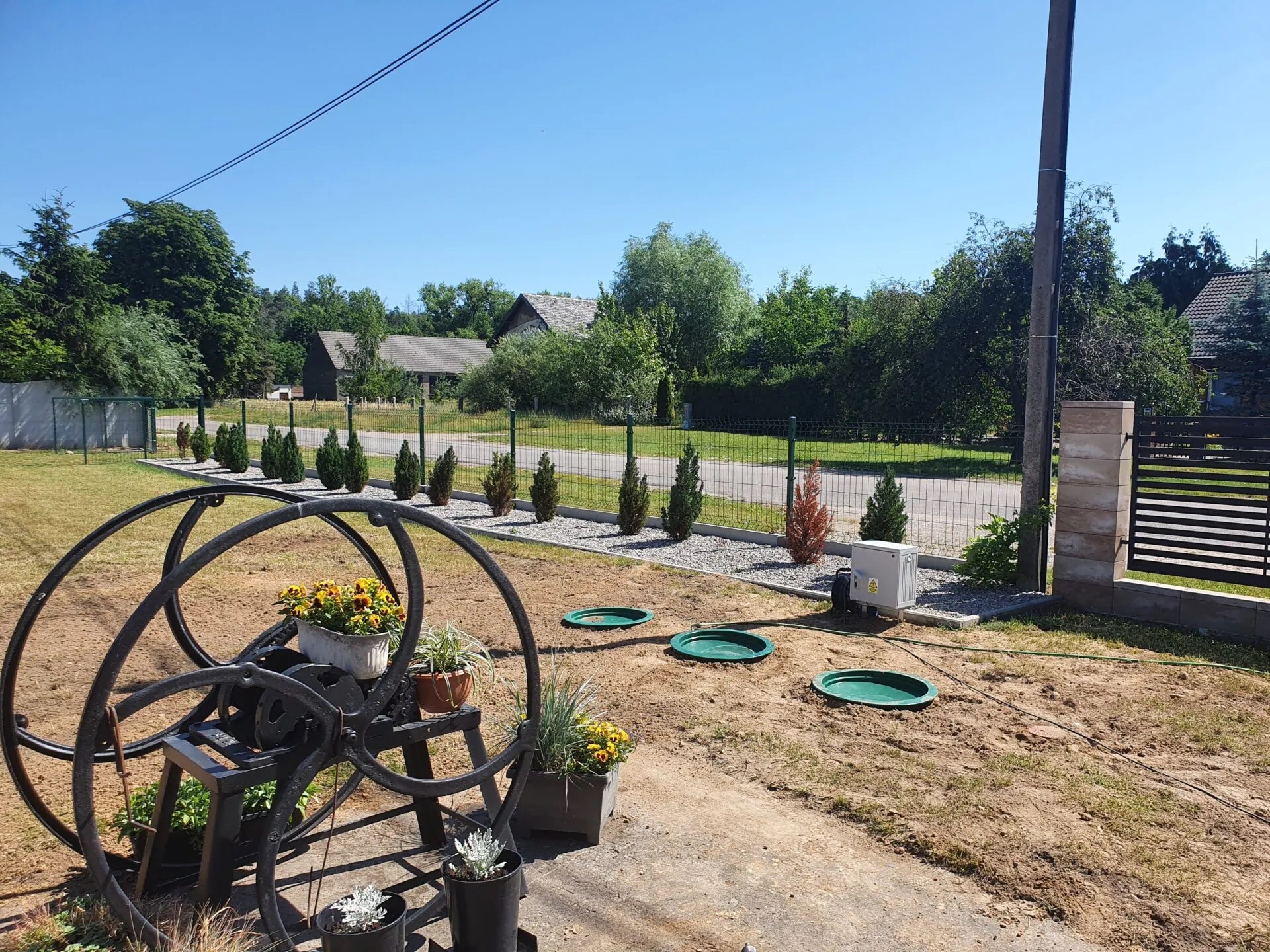 Widok na posesję z zainstalowaną przydomową oczyszczalnią ścieków z trzema zielonymi włazami, skrzynką sterującą i dekoracyjną konstrukcją z kwiatami na pierwszym planie.