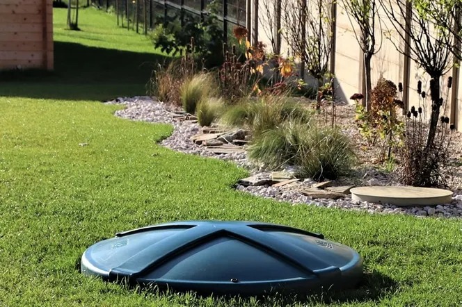 Zielony trawnik z okrągłą, ciemną pokrywą włazu do instalacji podziemnej, w tle ozdobne krzewy, kamienie i drewniana konstrukcja.