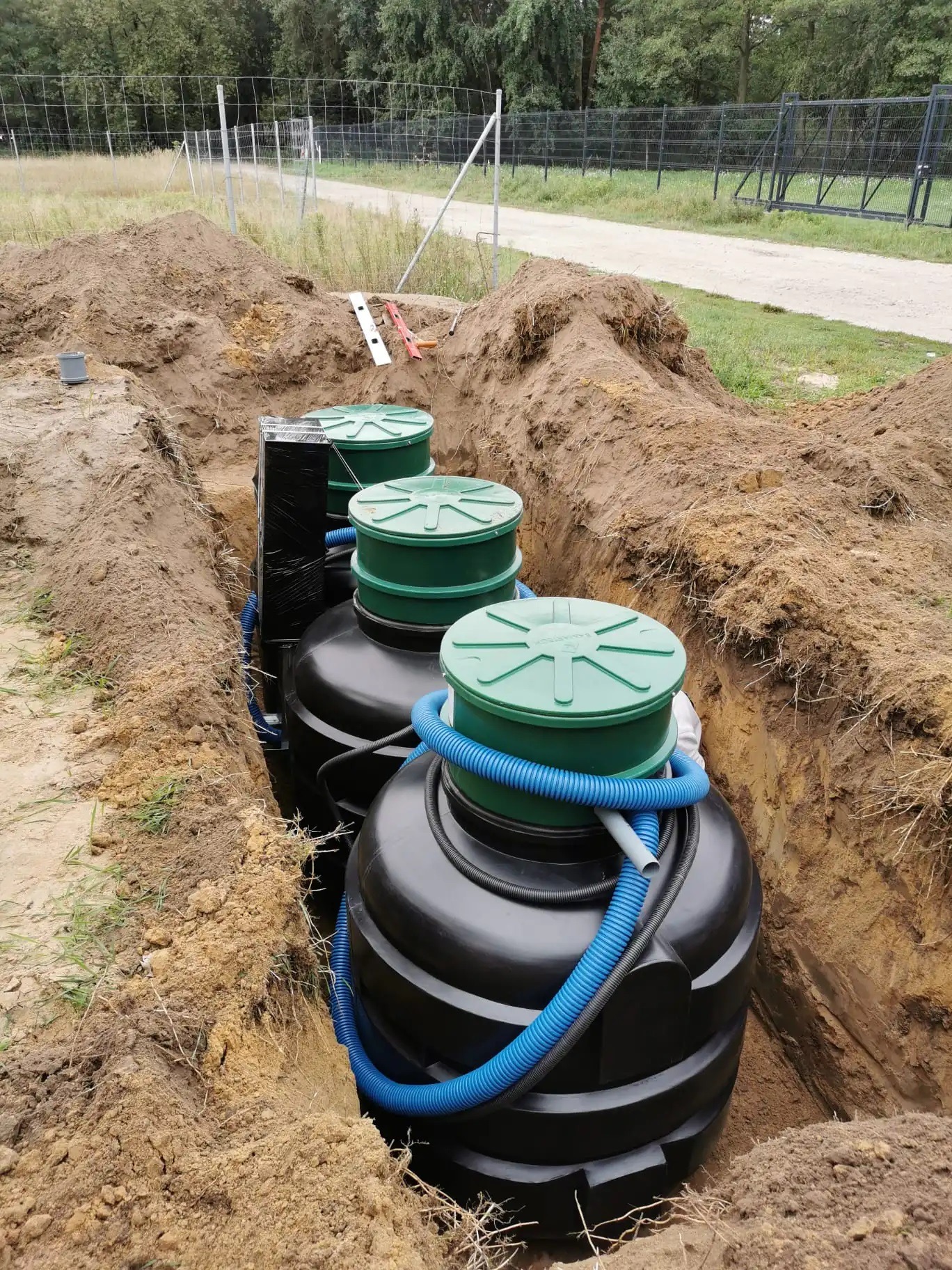 Instalacja trzech zbiorników z zielonymi pokrywami w wykopie, widoczne niebieskie rury karbowane i czarna folia ochronna, ziemia wokół, w tle ogrodzenie z siatki i droga gruntowa.