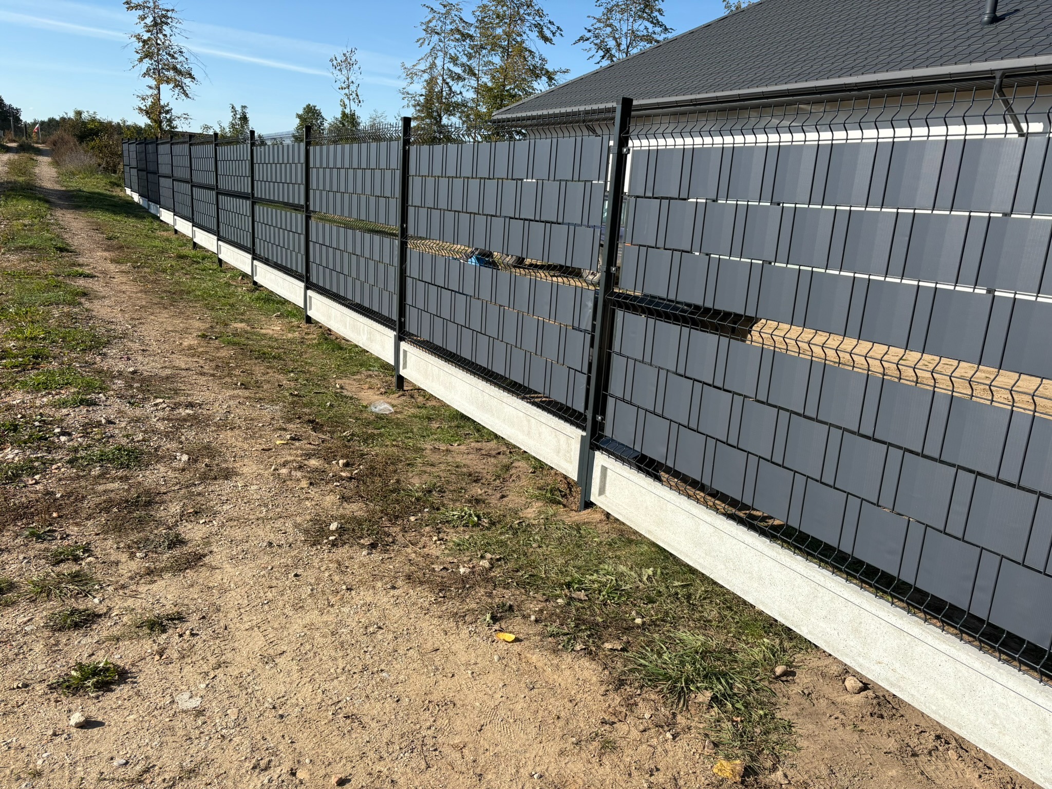 Nowoczesne ogrodzenie panelowe z szarymi wypełnieniami, osadzone na betonowej podmurówce, widoczne z perspektywy ścieżki gruntowej w słoneczny dzień.