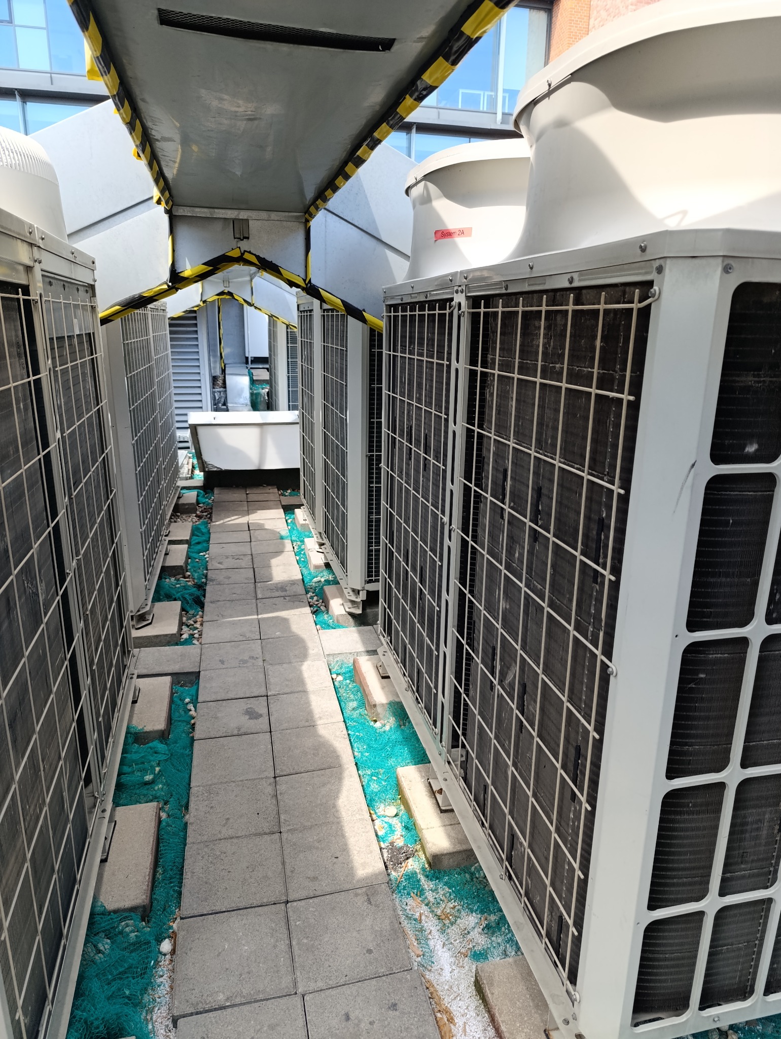 Rząd białych jednostek klimatyzacyjnych na dachu budynku, połączonych zadaszeniem z żółto-czarnymi pasami ostrzegawczymi, widoczne elementy instalacji i chodnik z betonowych płyt.