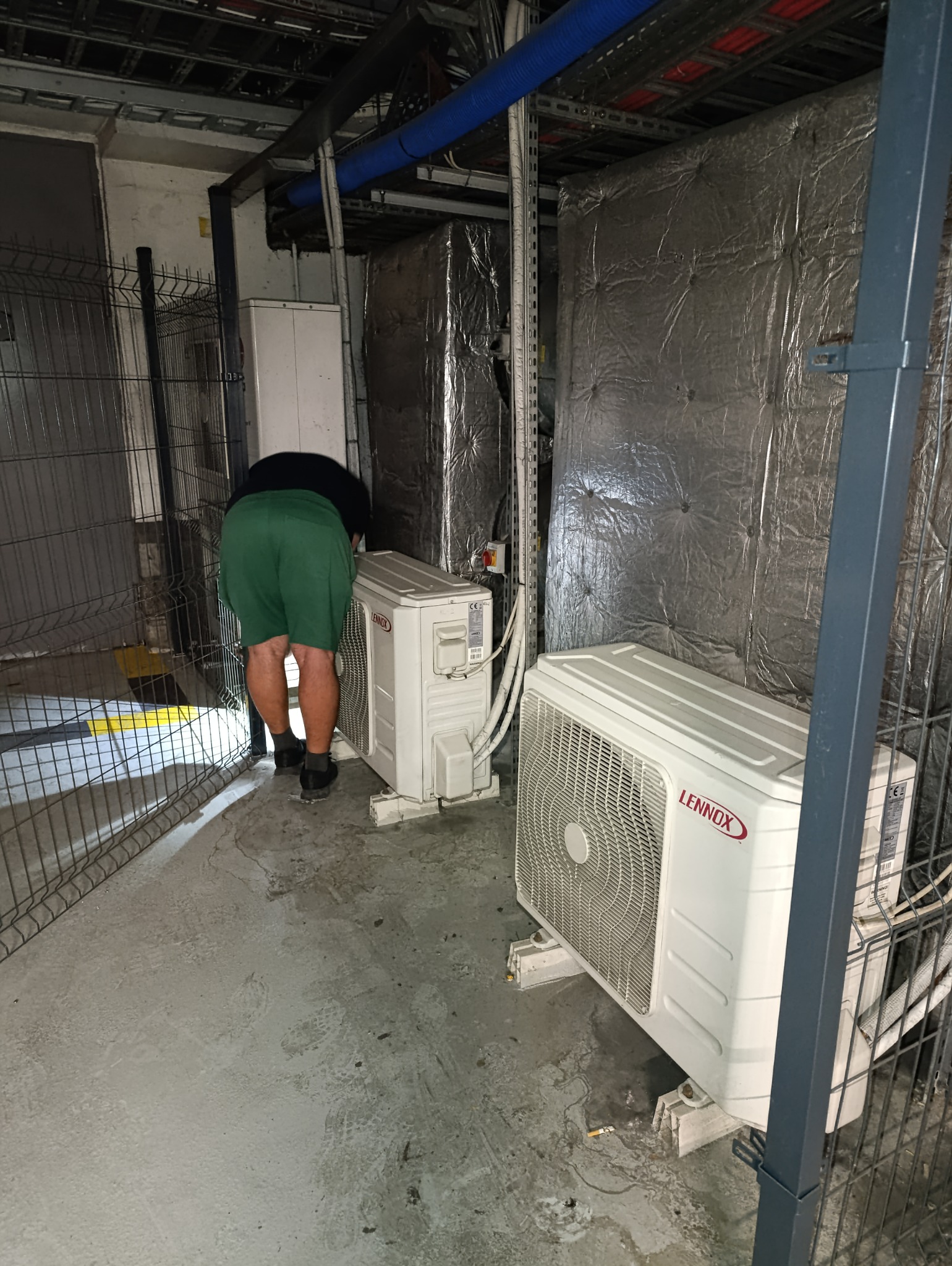 Instalacja dwóch klimatyzatorów marki Lennox w pomieszczeniu technicznym, widoczny pracownik w zielonych szortach pochylony nad jednym z urządzeń za metalową siatką, w tle izolowane kanały...