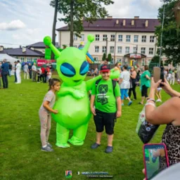 Dzieci pozują z animatorem w kostiumie zielonego kosmity na trawniku podczas imprezy plenerowej. W tle budynek i inne osoby.