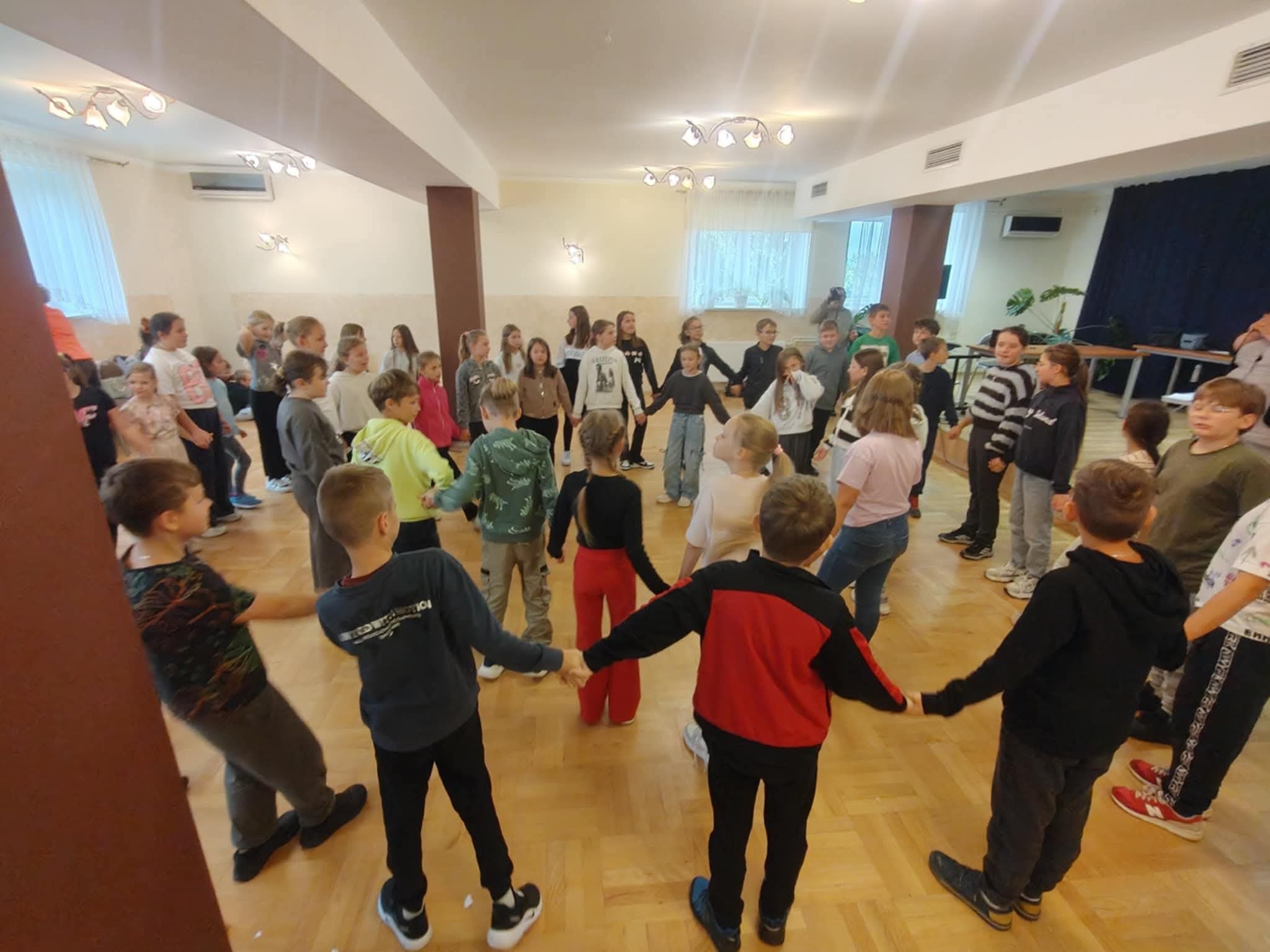 Dzieci trzymają się za ręce, tworząc koło w jasnej sali. Uczestnicy w różnorodnych strojach, drewniana podłoga i kolumny w tle. Zabawa z animatorem.