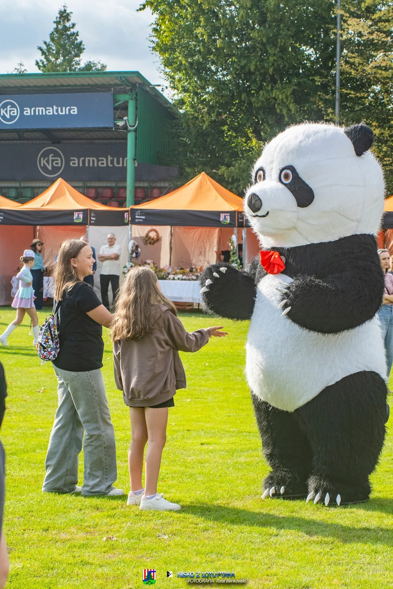 Dwie dziewczynki witają się z animatorem w kostiumie pandy z czerwoną muchą na tle zielonego trawnika i rozstawionych namiotów.