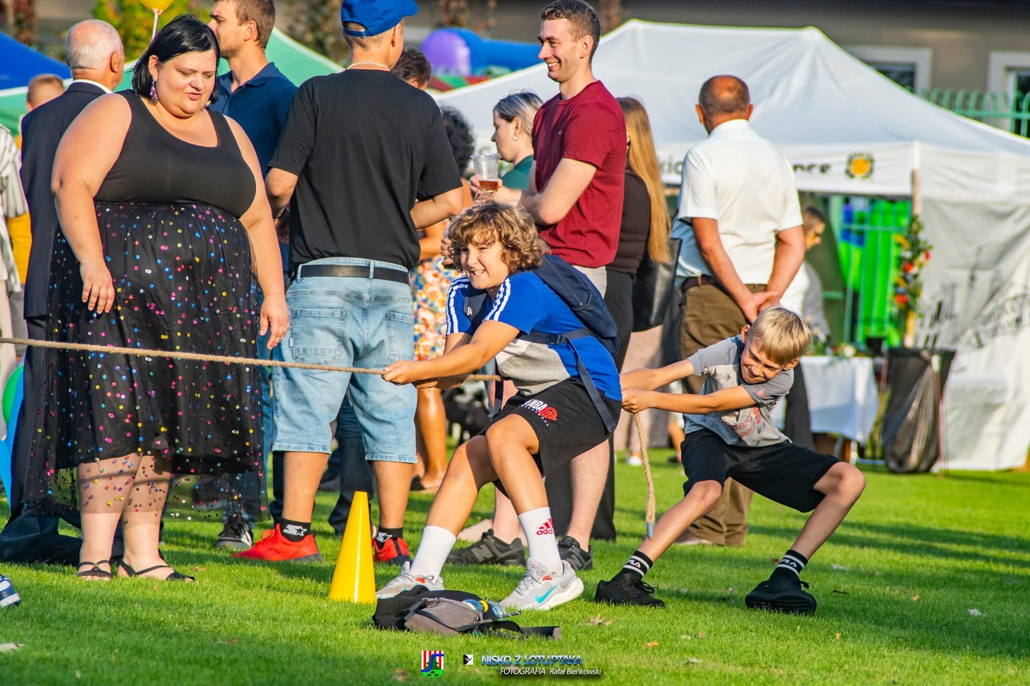 Dzieci biorące udział w przeciąganiu liny na trawniku podczas festynu. W tle widoczni dorośli i biały namiot.