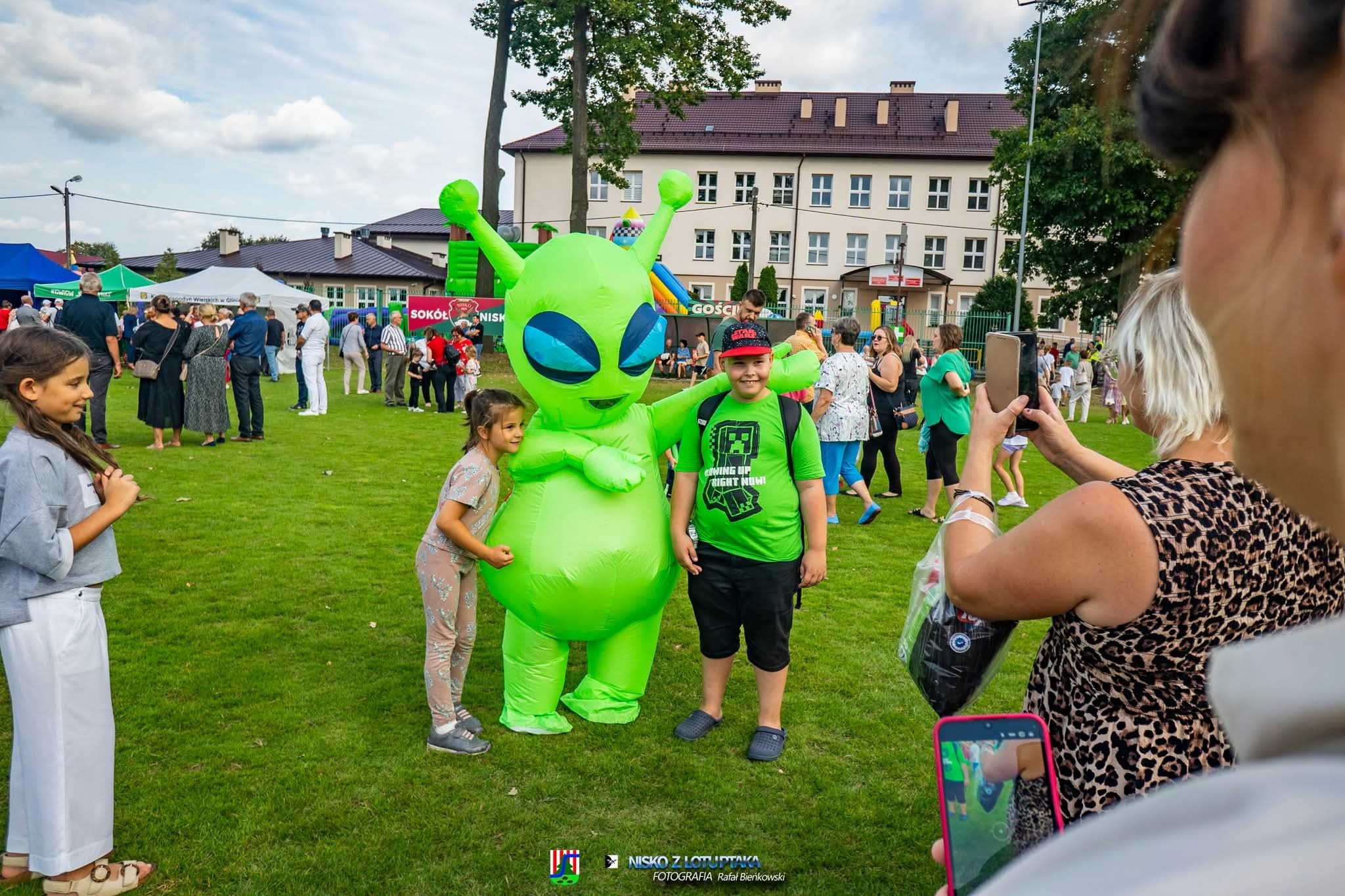 Dzieci pozują z animatorem w kostiumie zielonego kosmity na trawniku podczas imprezy plenerowej. W tle budynek i inne osoby.