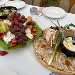 Elegancko podane deski z serami, owocami i wędlinami na białym obrusie, z zielonymi dodatkami. Widoczne naczynia i szklanki, sugerujące gotowość do spożycia.