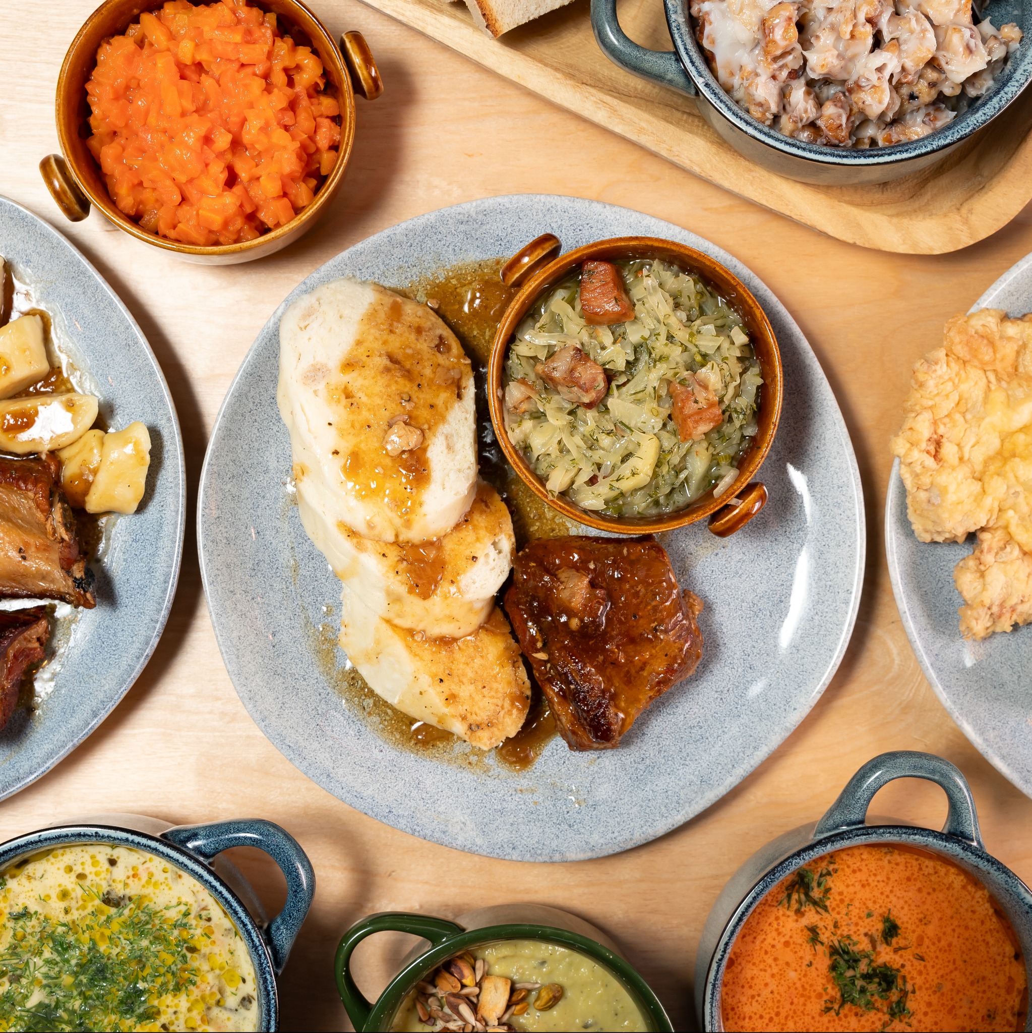 Zestaw dań obiadowych na drewnianym stole: kluski śląskie z sosem, kapusta zasmażana z boczkiem w brązowym naczyniu, pieczeń, surówka z marchwi, zupa krem z zielonymi przyprawami, zupa krem...