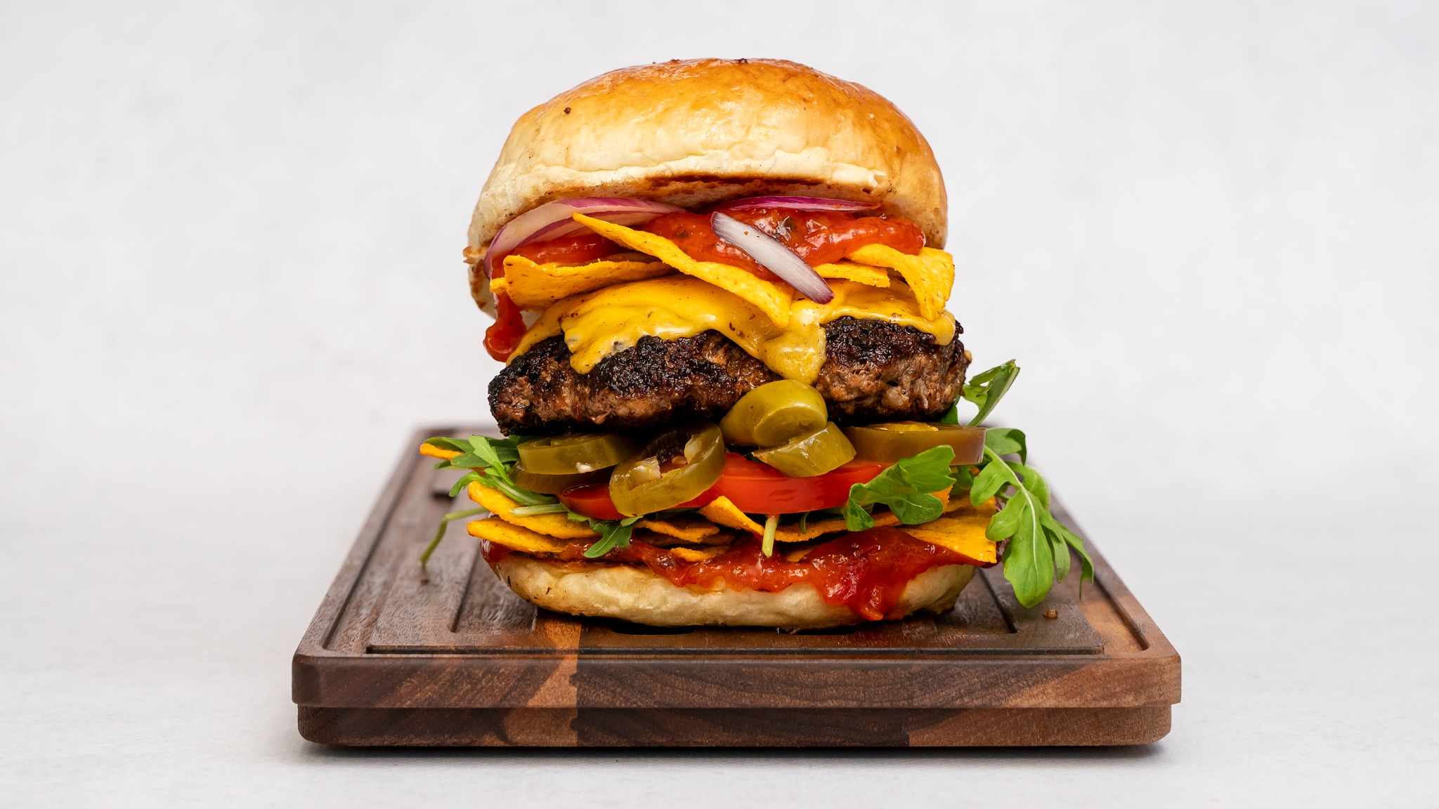 Apetyczny, wysoki burger z wołowiną, nachosami, serem cheddar, czerwoną cebulą, sosem pomidorowym, jalapeno i rukolą, ułożony na drewnianej desce.