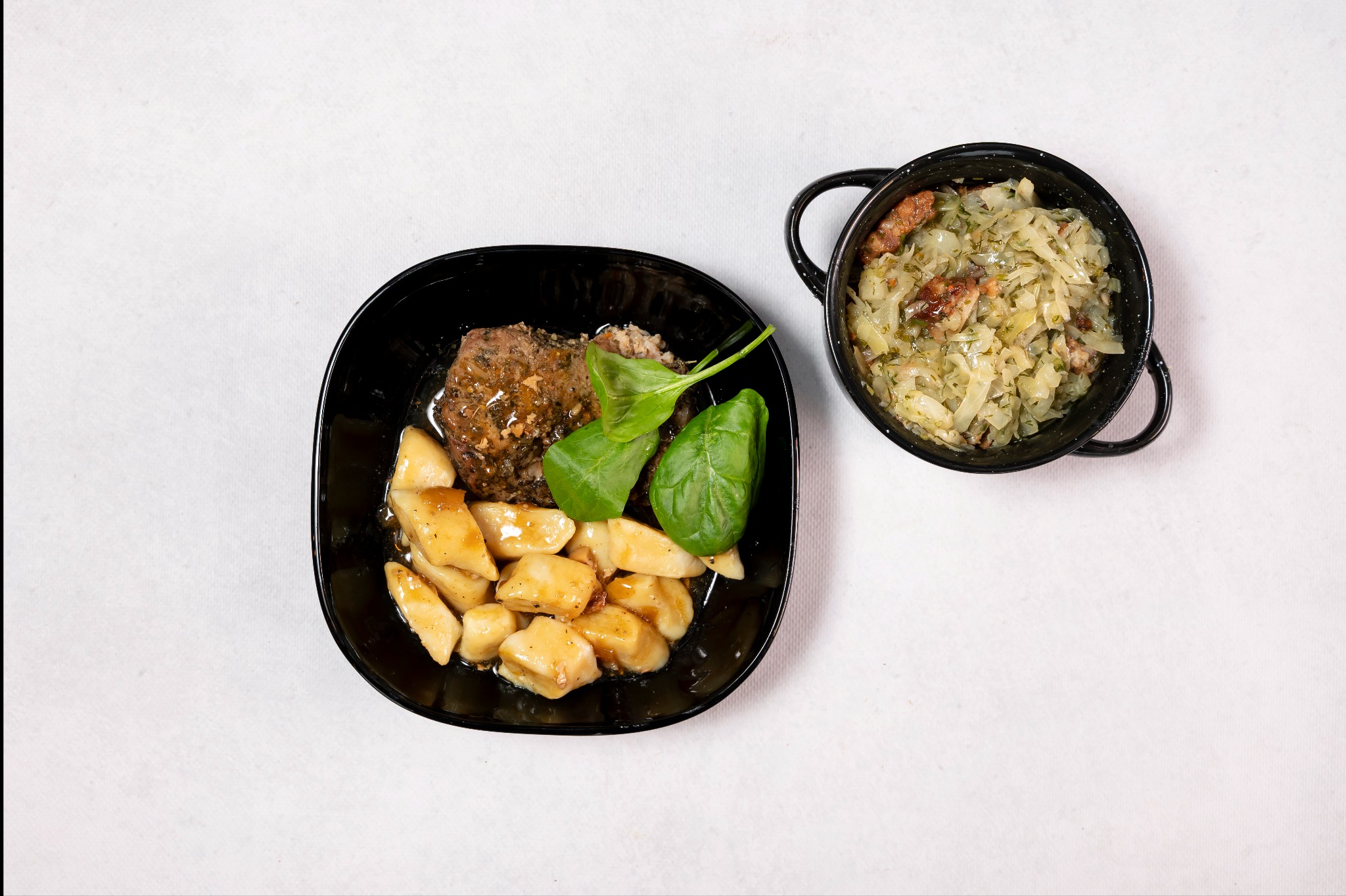 Danie obiadowe: kluski, mięso w sosie i liście szpinaku na czarnym talerzu oraz kapusta zasmażana w czarnym garnku z uchwytami, widok z góry na białym tle.