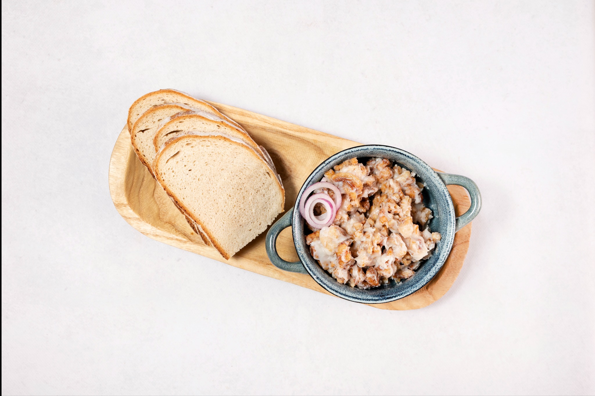 Smalec ze skwarkami w niebieskiej miseczce z uchwytami, udekorowany krążkami czerwonej cebuli, podany z kromkami chleba na drewnianej desce.