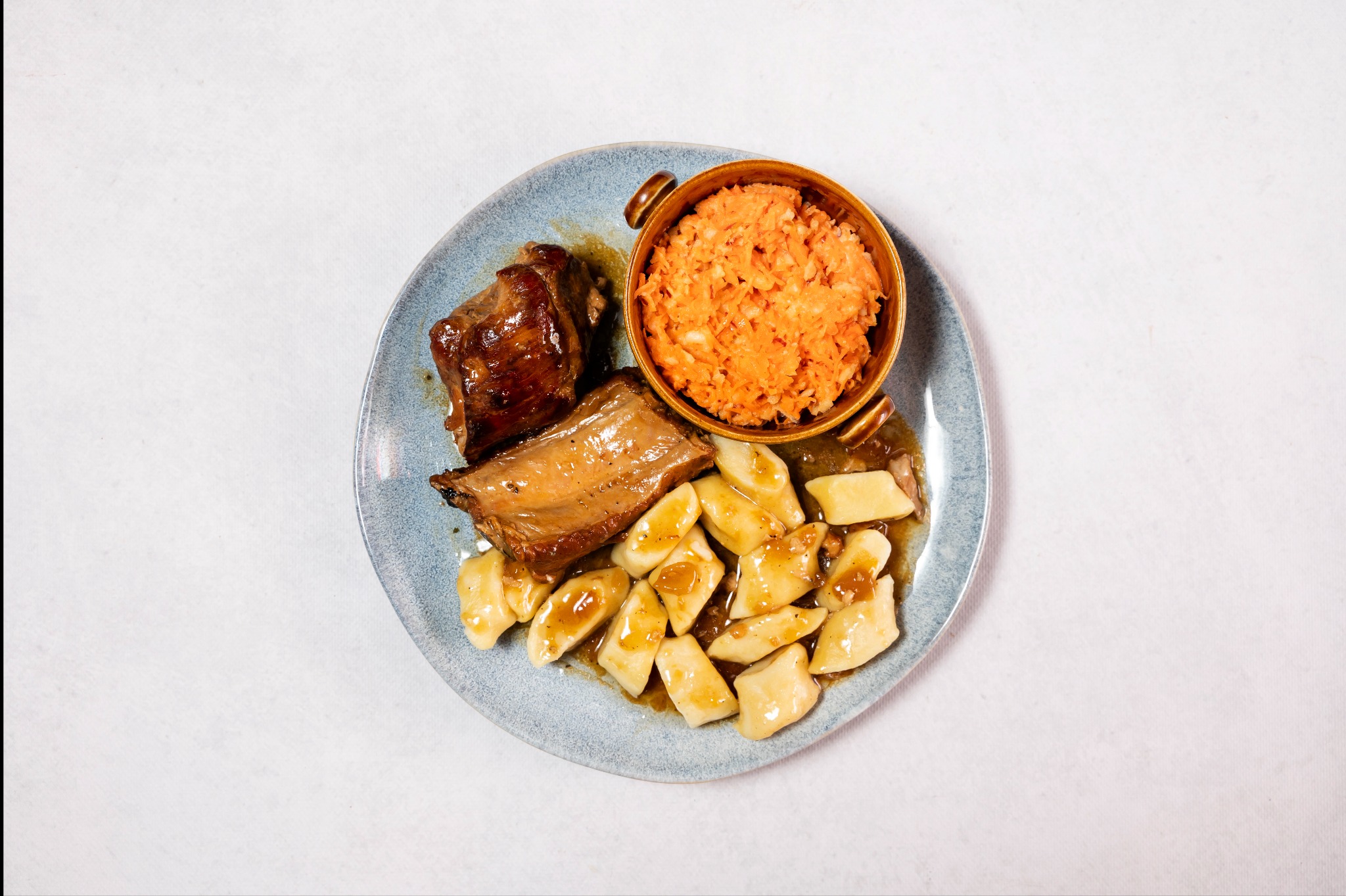 Danie główne na niebieskim talerzu: dwa kawałki mięsa w sosie, kluski oraz marchewka tarta w małym garnuszku.