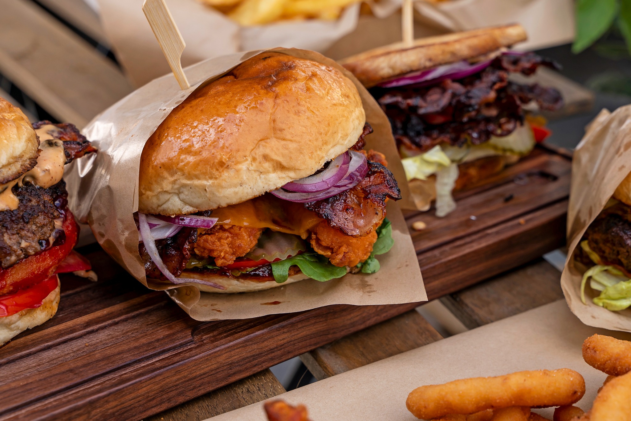 Ręcznie robione burgery z kurczakiem, bekonem, czerwoną cebulą i sosem, podane na drewnianej desce z frytkami, zapakowane w papier.