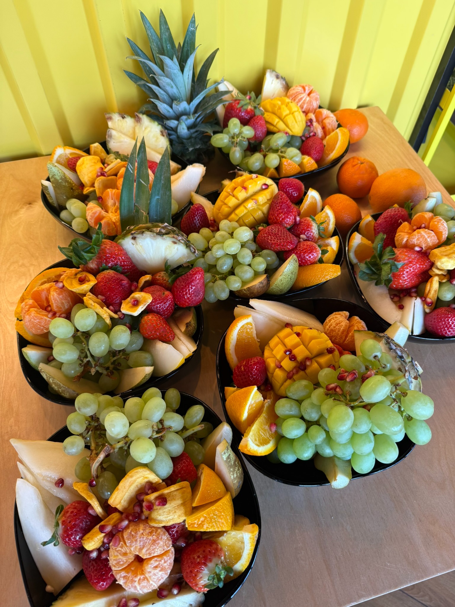 Egzotyczne owoce: mango, ananas, truskawki, winogrona i pomarańcze, artystycznie ułożone w czarnych misach na cateringowym stole z żółtym tłem.