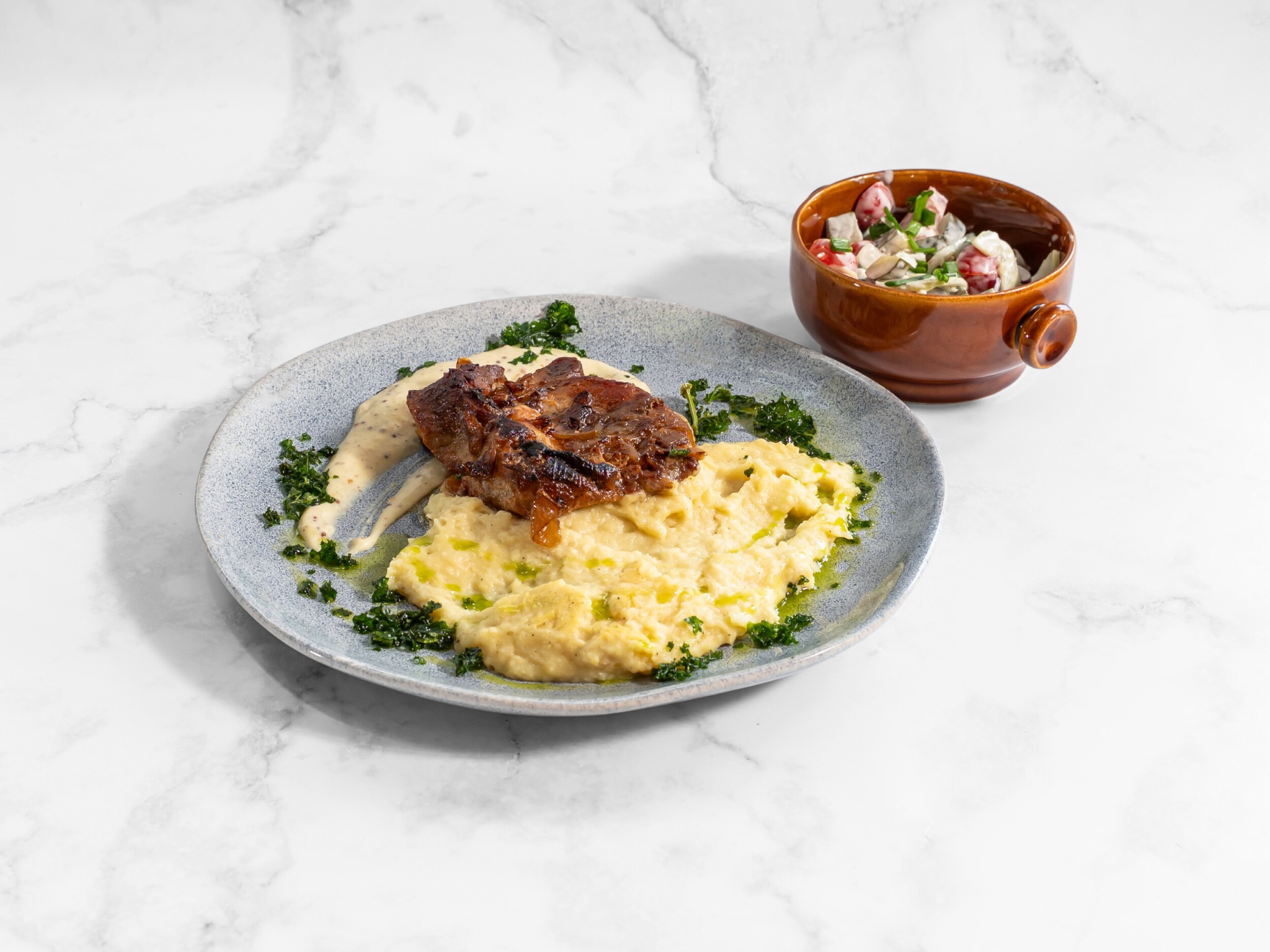 Elegancko podane danie: mięso na puree z sosem, ozdobione zielenią, z sałatką w brązowej miseczce na marmurowym tle, idealne na catering.