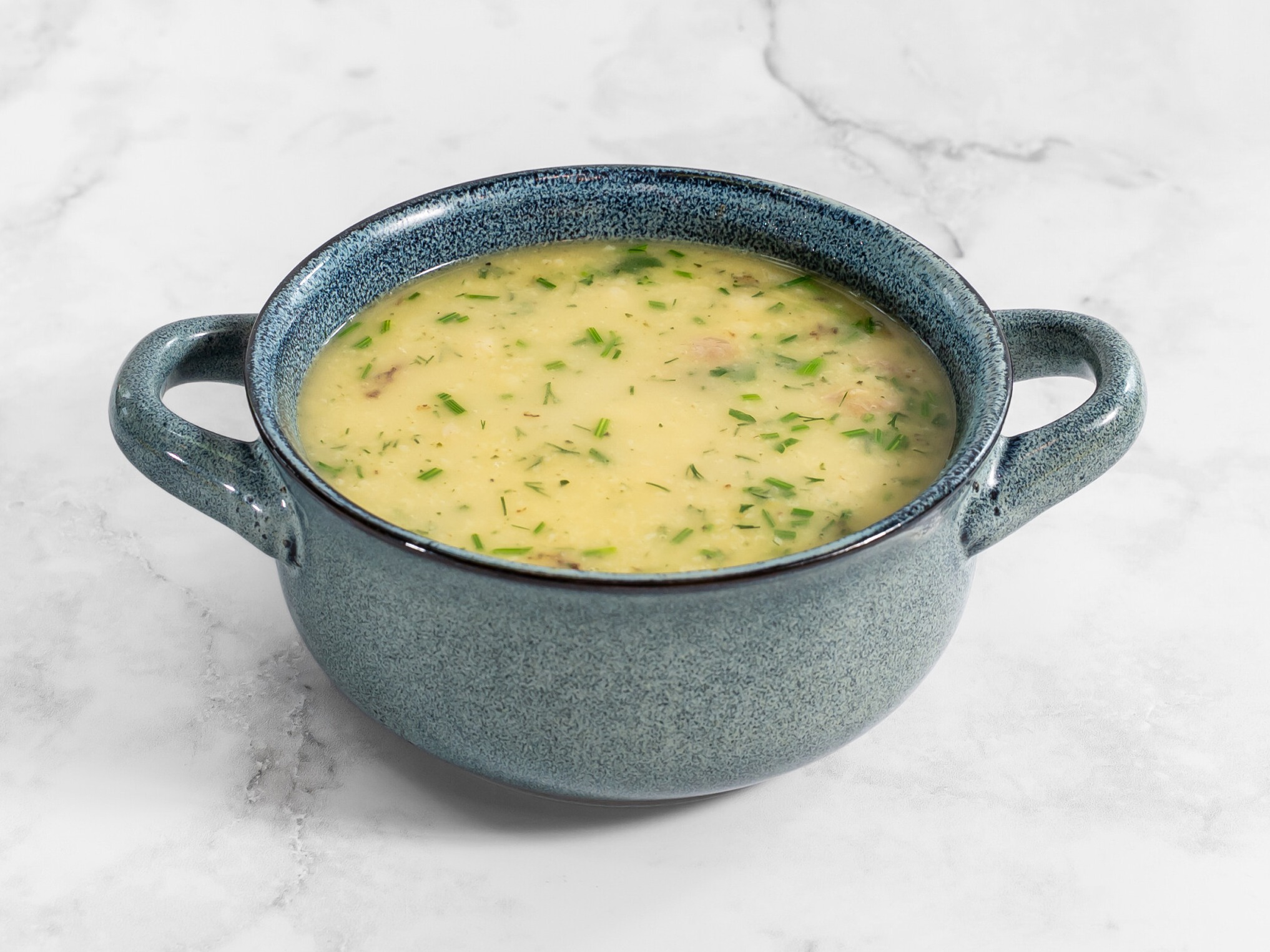 Kremowa zupa w niebieskiej miseczce z uchwytami, posypana świeżym koperkiem, na białym marmurowym tle. Delikatne oświetlenie podkreśla fakturę naczynia.