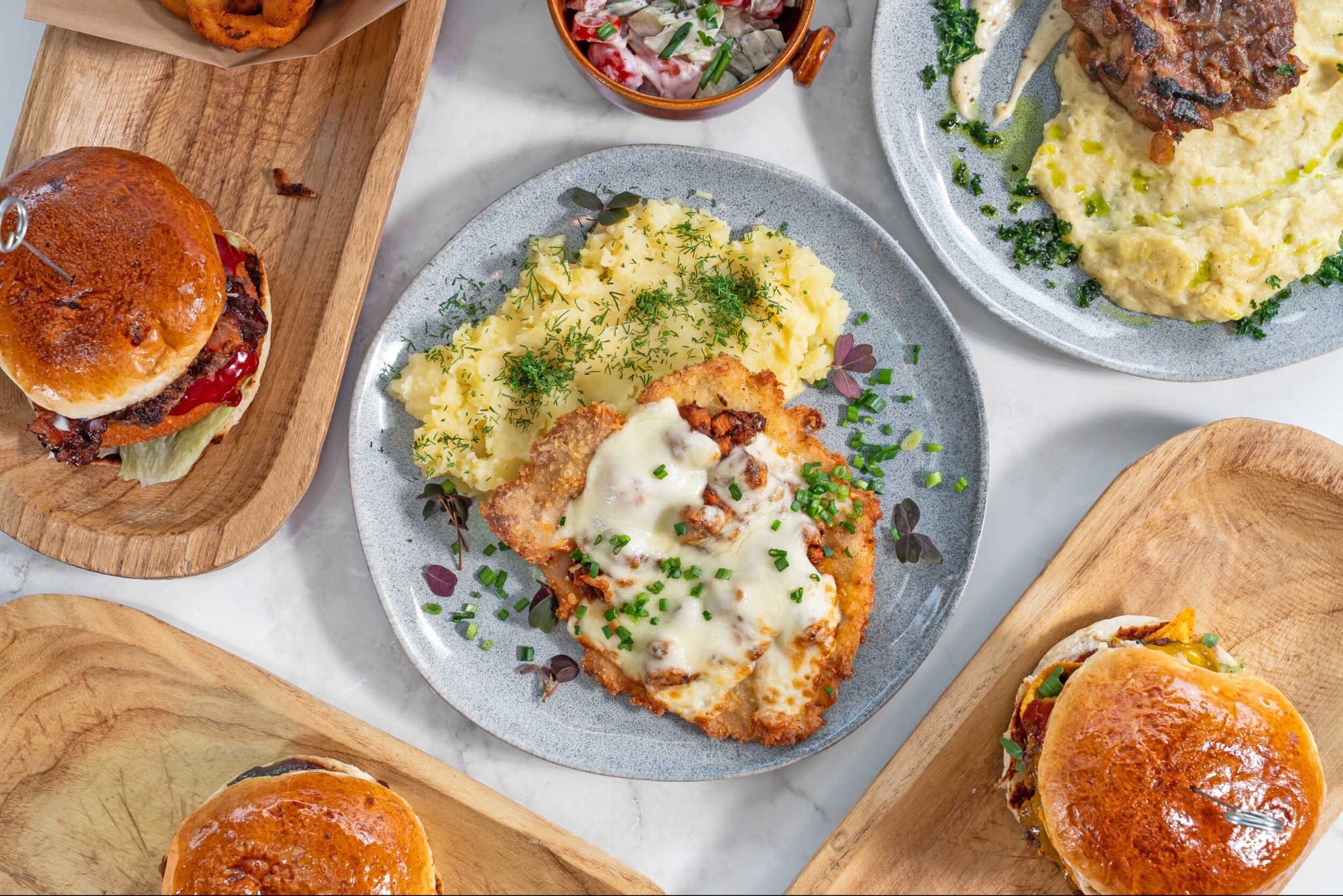 Zestaw dań: kotlet z serem i puree na talerzu, burger na desce, sałatka w miseczce, stek z puree na talerzu. Jasne, naturalne światło, widok z góry.