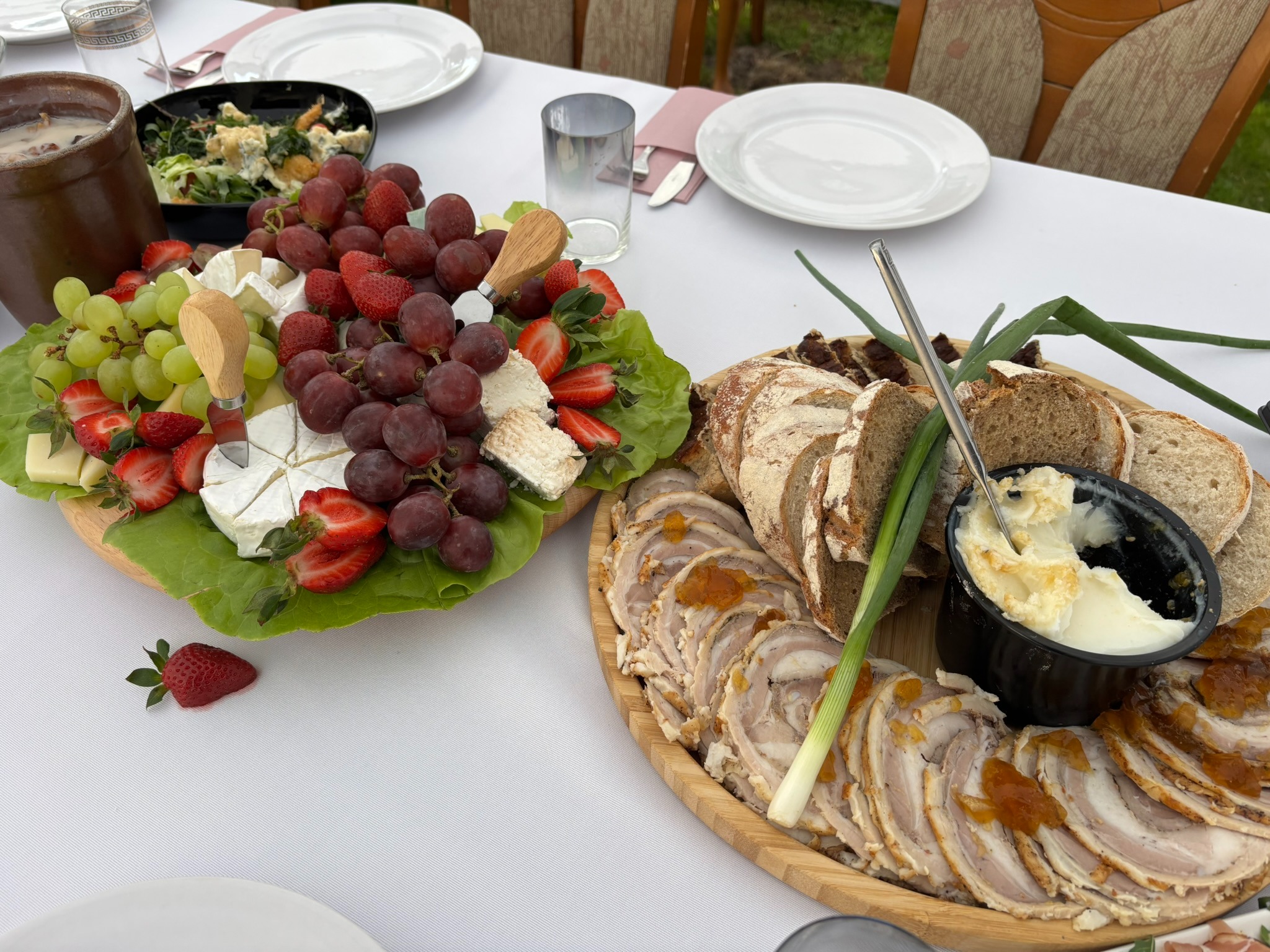 Elegancko podane deski z serami, owocami i wędlinami na białym obrusie, z zielonymi dodatkami. Widoczne naczynia i szklanki, sugerujące gotowość do spożycia.