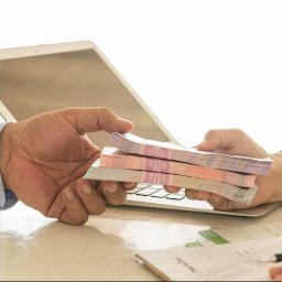 Social Financing Agency - Wymiana gotówki między dwiema osobami na tle laptopa i dokumentów, transakcja finansowa w biurze.